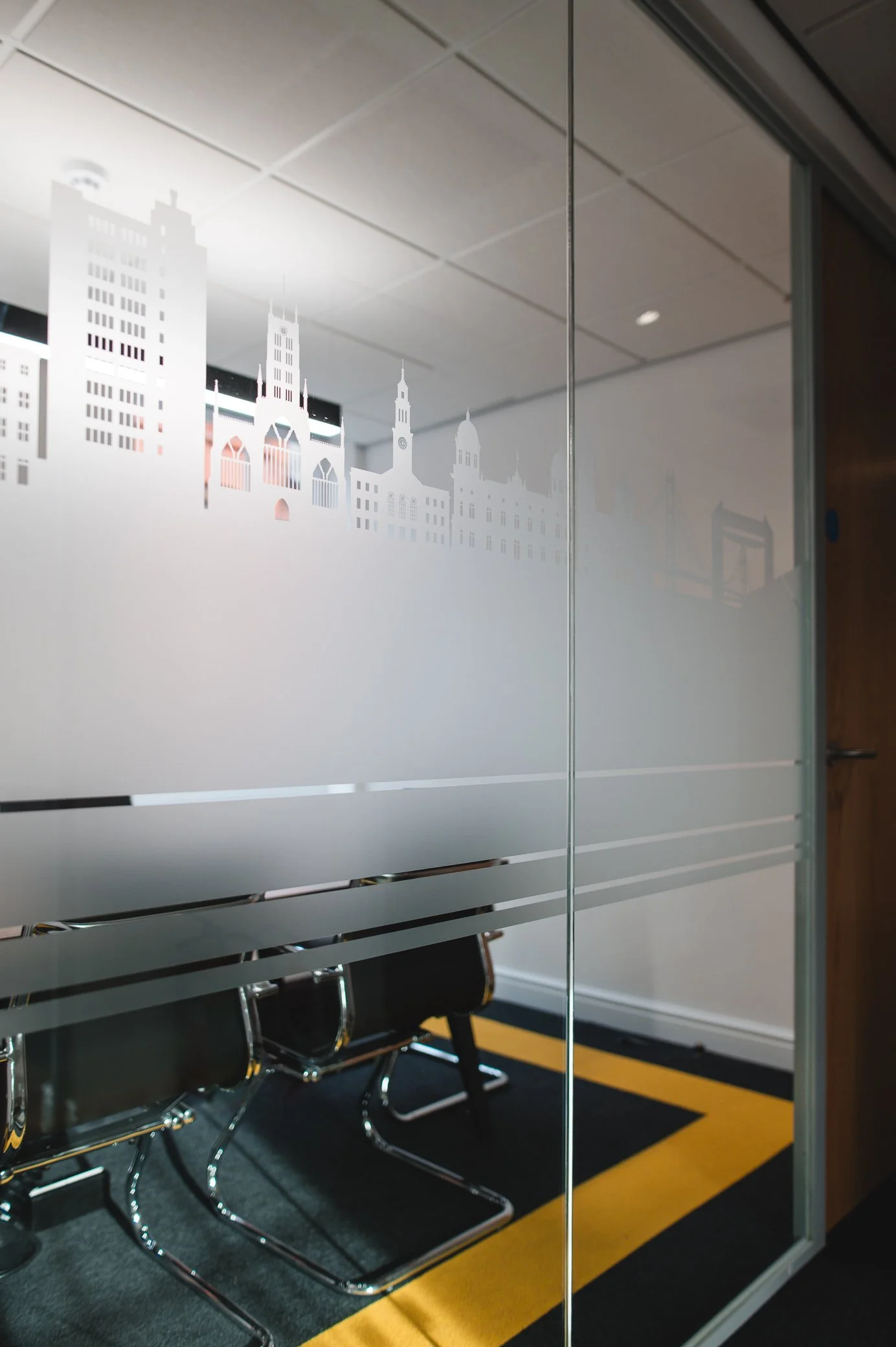 Interior of a conference room with black chairs, a decorative cityscape frosted glass wall, and a ceiling with recessed lighting.