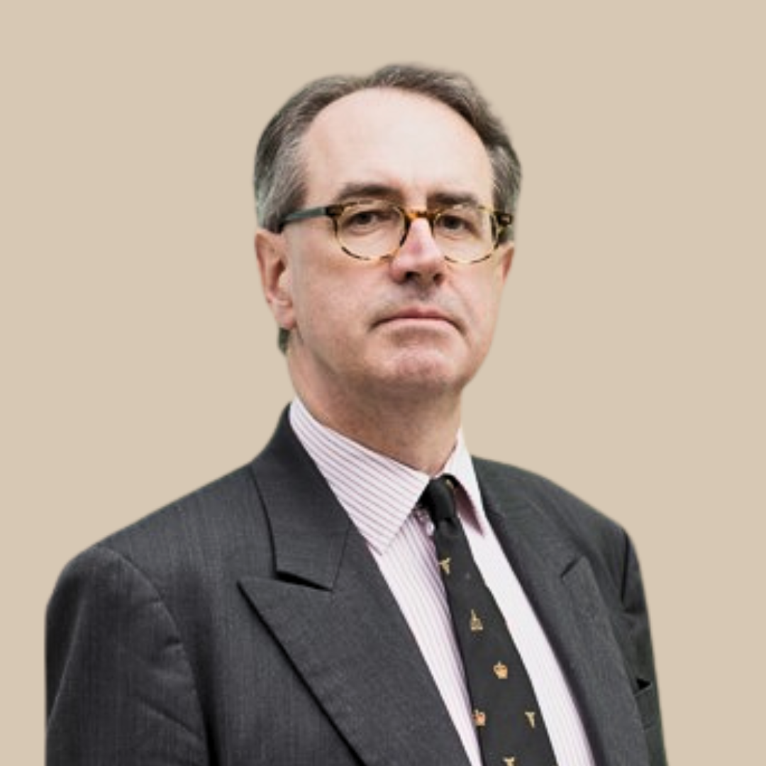 A middle-aged man wearing glasses, a dark suit, a striped dress shirt, and a black tie with small yellow and white icons, standing against a beige background.