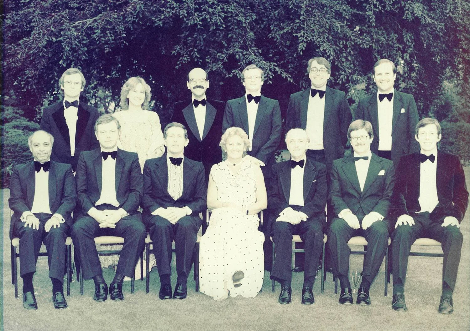 Group of fifteen people dressed in formal tuxedos and a dress, posing outdoors in front of trees. The group is arranged in two rows, with some seated and others standing.