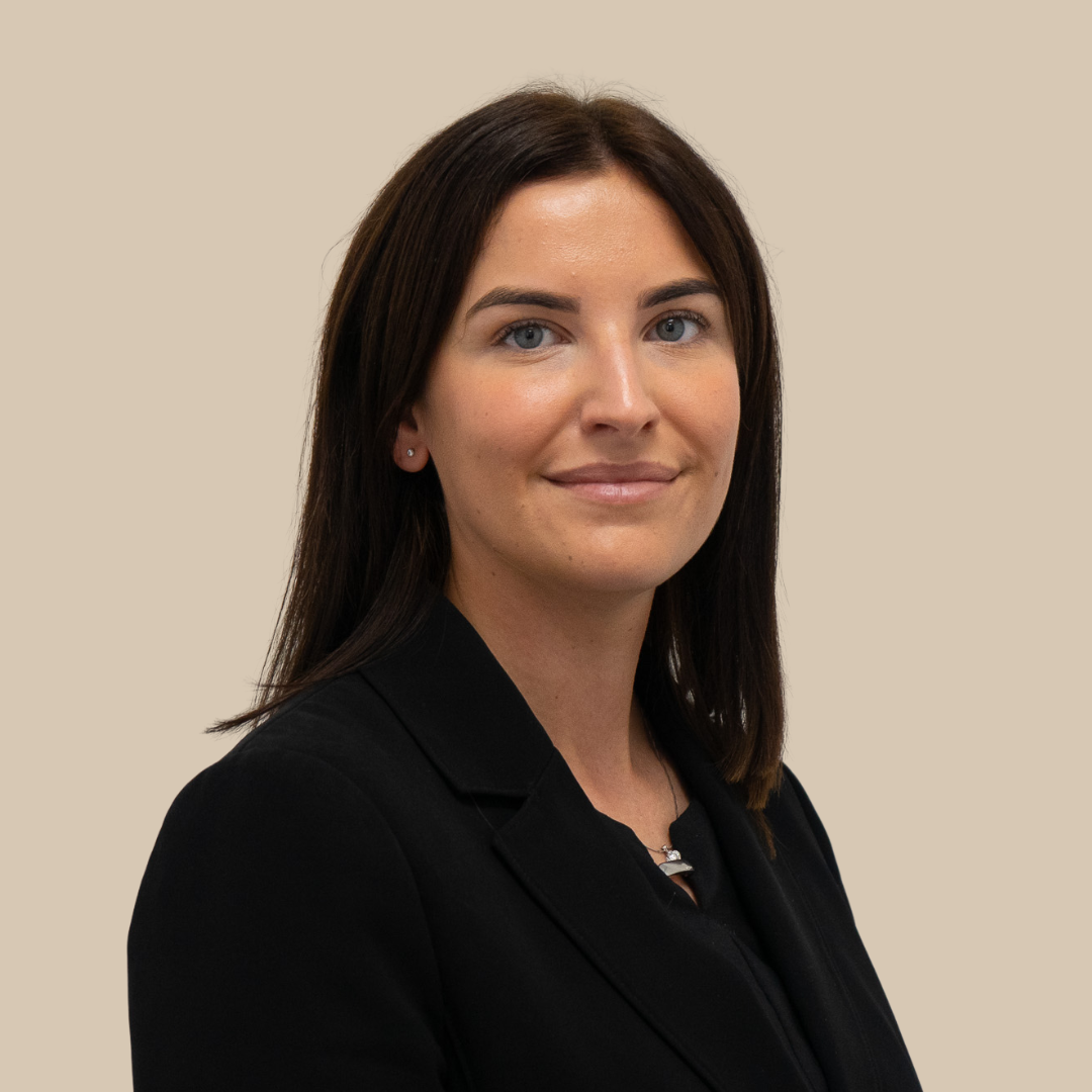 A woman with dark brown hair, blue eyes, and fair skin, wearing a black blazer, smiling slightly, against a beige background.