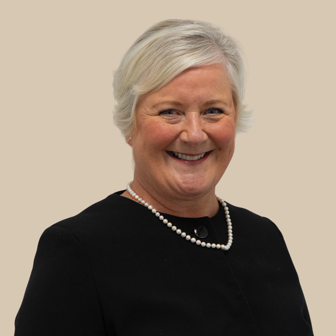 Portrait of a smiling woman with short white hair wearing a black top and pearl necklace, against a beige background.