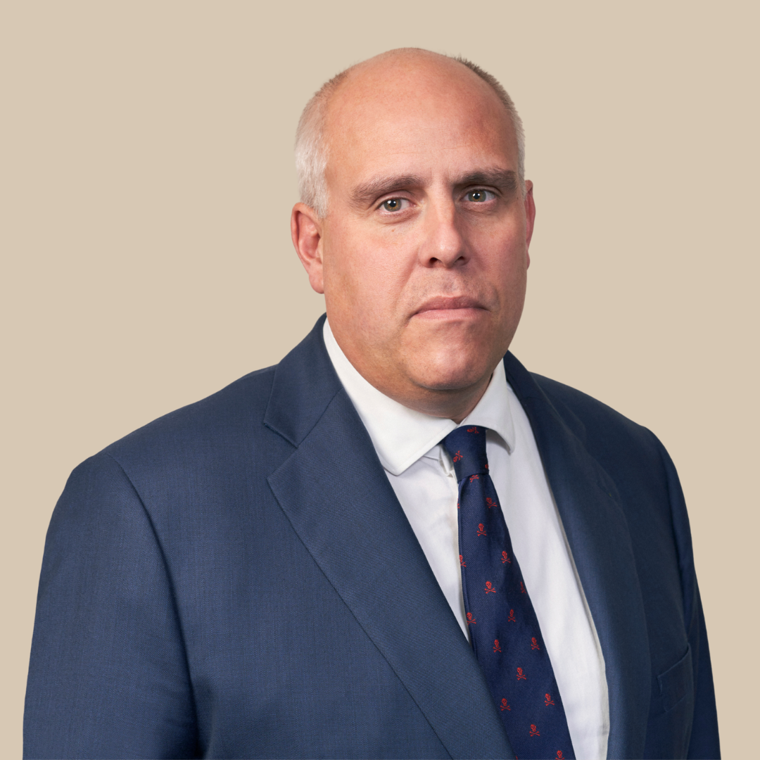 A middle-aged man with a serious expression, dressed in a navy blue suit, white shirt, and a navy tie with small red patterns, against a light beige background.