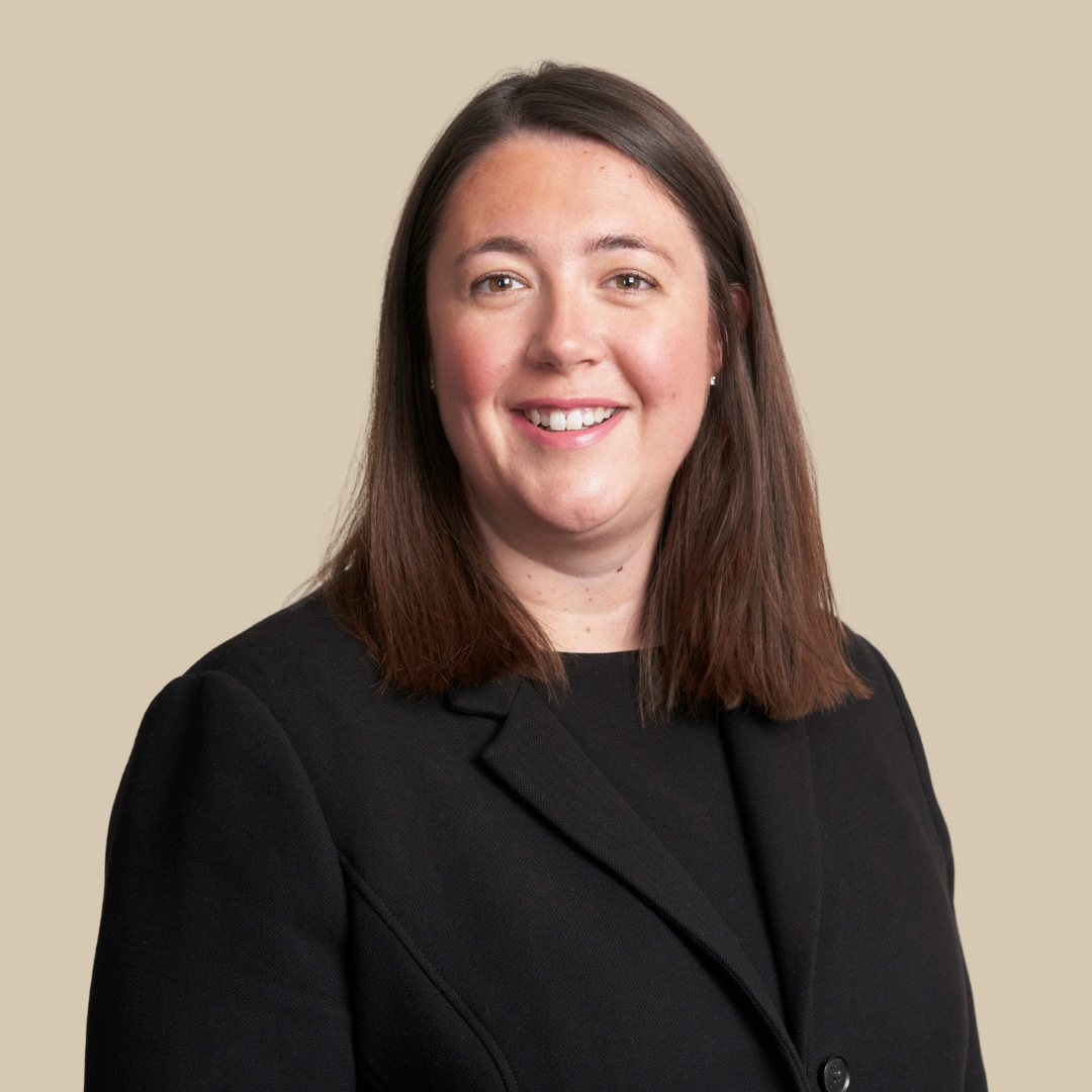 A woman with shoulder-length brown hair, smiling, wearing a black blazer and a black top, against a beige background.