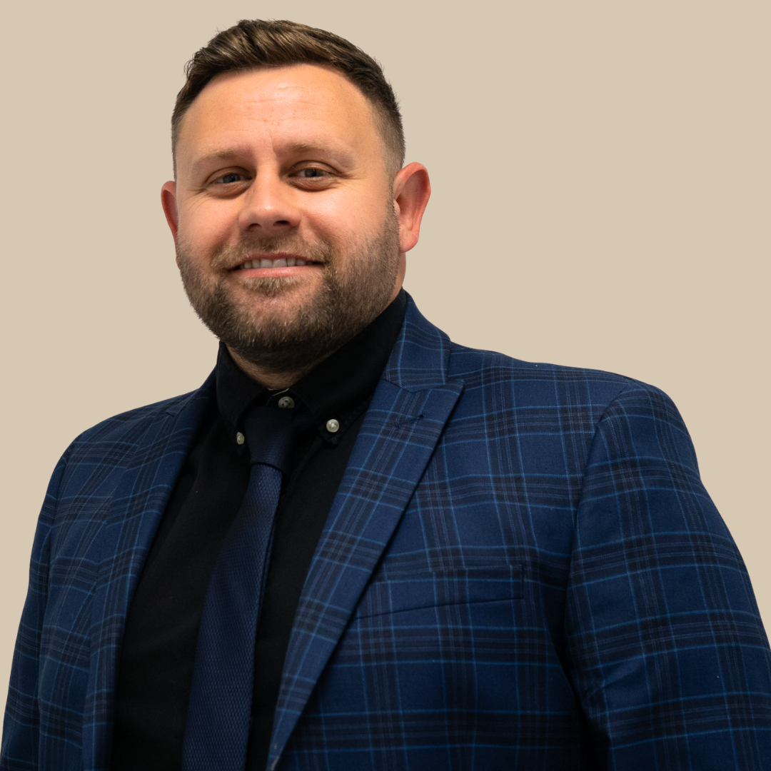 Portrait of a man with a beard and short hair, wearing a dark blue plaid blazer and a dark shirt with a tie, smiling against a plain beige background.