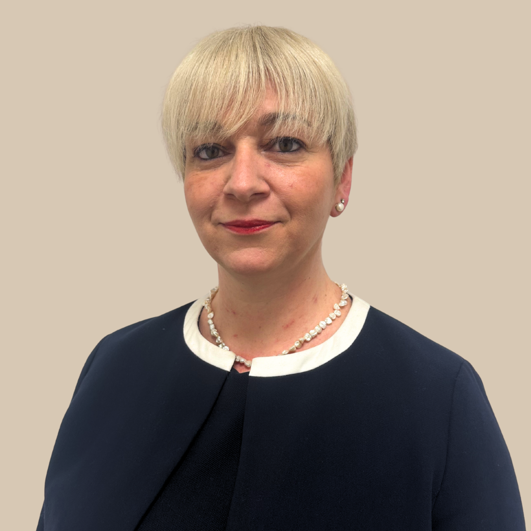 A woman with short, blonde hair wearing a navy blue blazer with white trim, pearl earrings, a pearl necklace, and red lipstick, standing against a plain beige background.