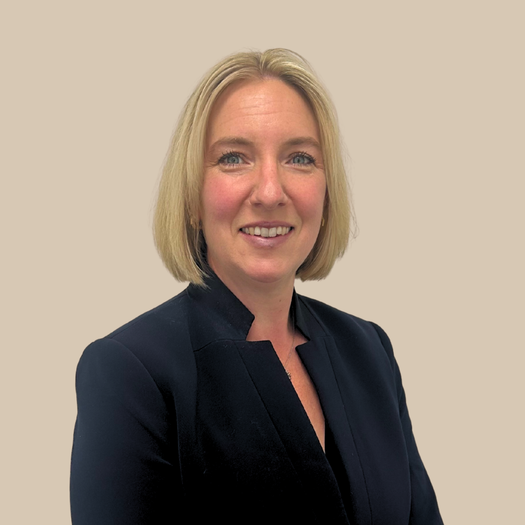 A woman with shoulder-length blonde hair, wearing a black blazer, smiling against a plain beige background.