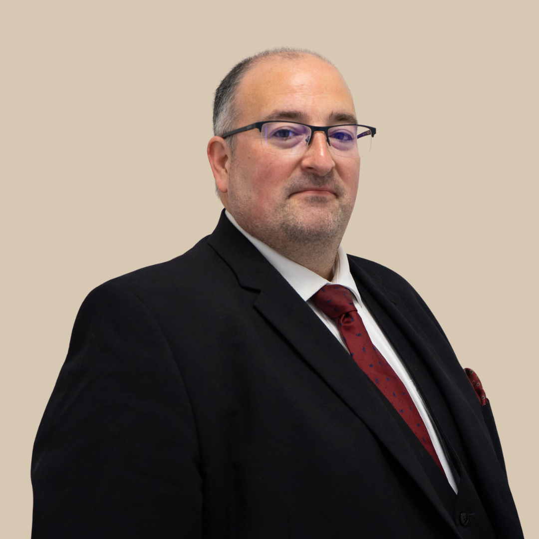 A middle-aged man with glasses, dressed in a black suit, white shirt, and red tie, standing against a beige background.