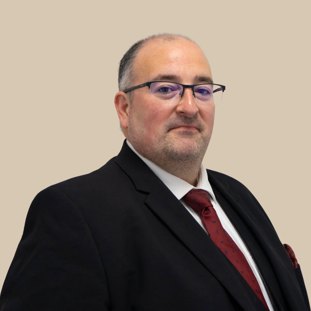 A man in a black suit with a white shirt and red tie, wearing glasses, posing against a plain beige background.