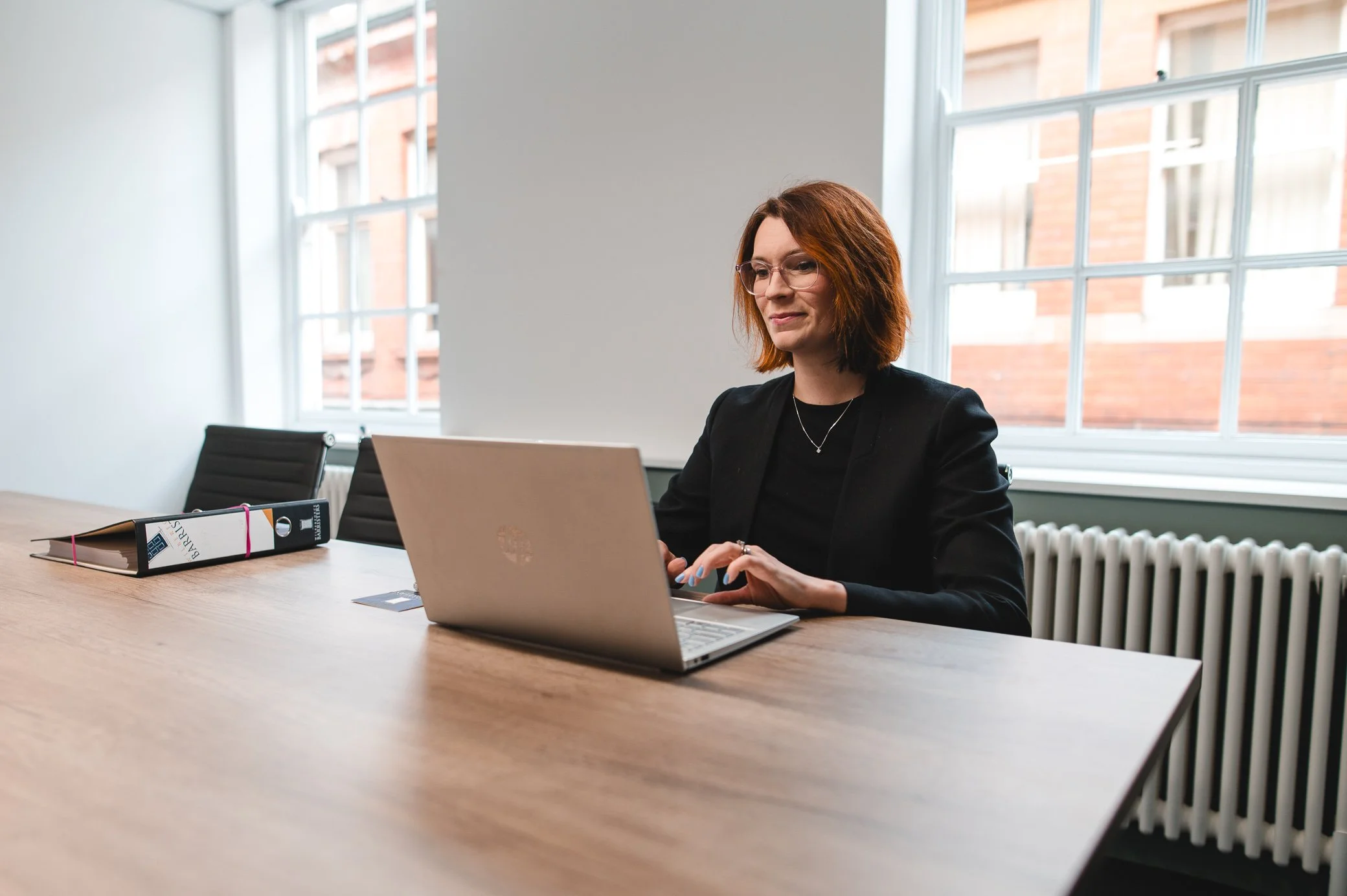A woman with red hair and glasses working on a laptop in a modern office with large windows and a radiator.