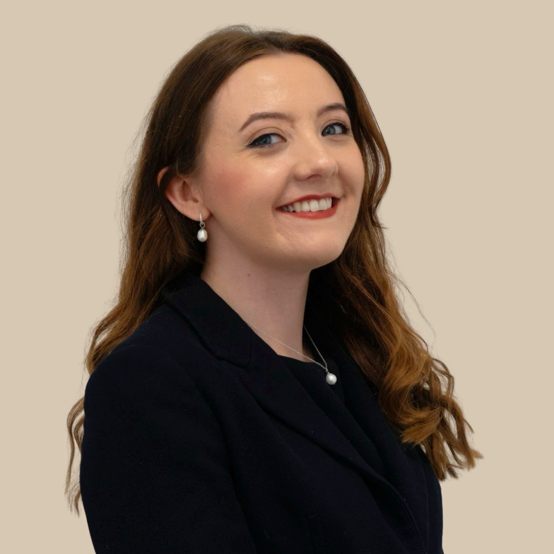 A portrait of a woman with long, wavy brown hair wearing a black blazer and pearl jewelry, smiling against a plain beige background.