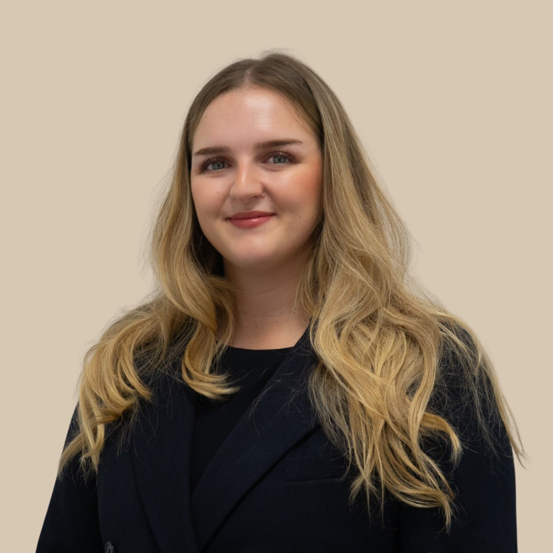 A young woman with long blonde wavy hair wearing a black blazer and top, smiling gently, against a beige background.