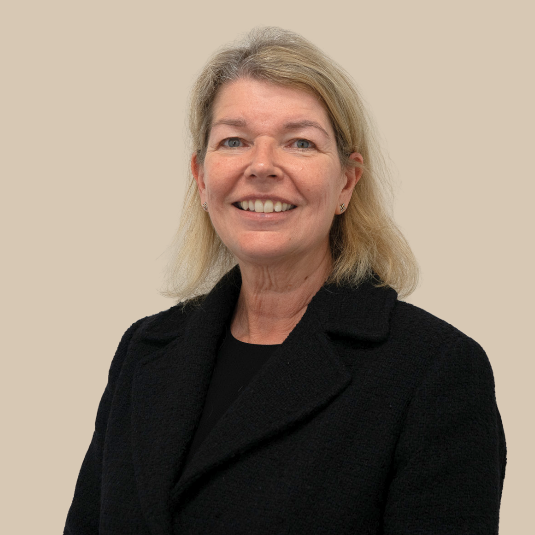 A middle-aged woman with shoulder-length blonde hair, wearing a black top and necklace, looking directly at the camera with a neutral expression, standing against a plain beige background.