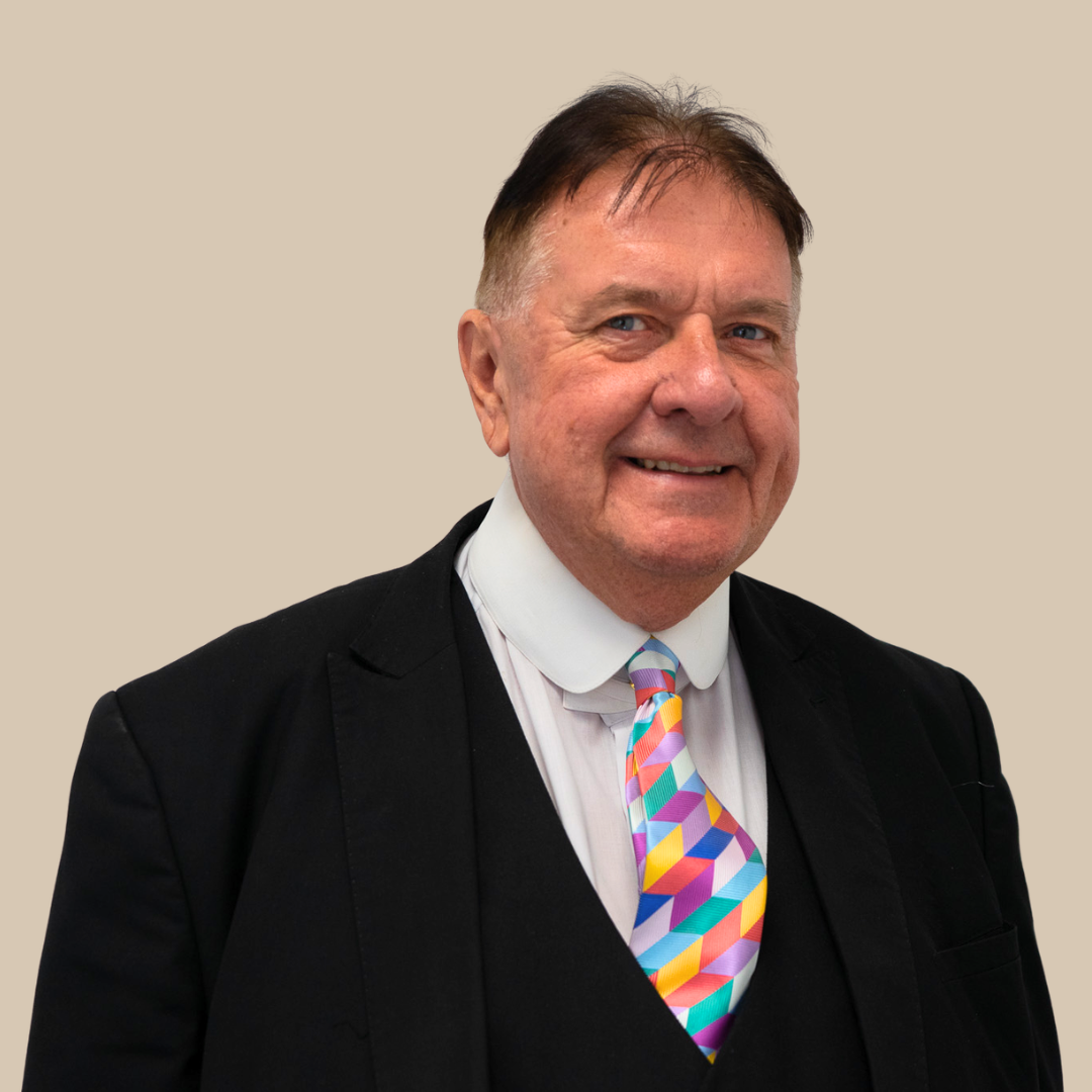 A man in a black suit with a white shirt and yellow striped tie, standing against a plain beige background.