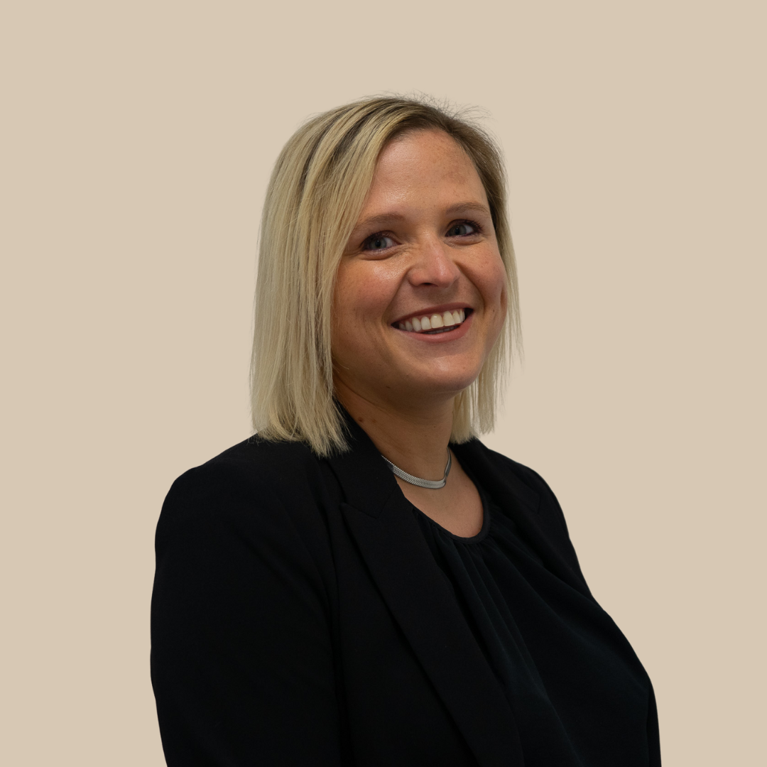 A smiling blonde woman with shoulder-length hair, wearing a black blazer and a silver necklace, standing against a beige background.