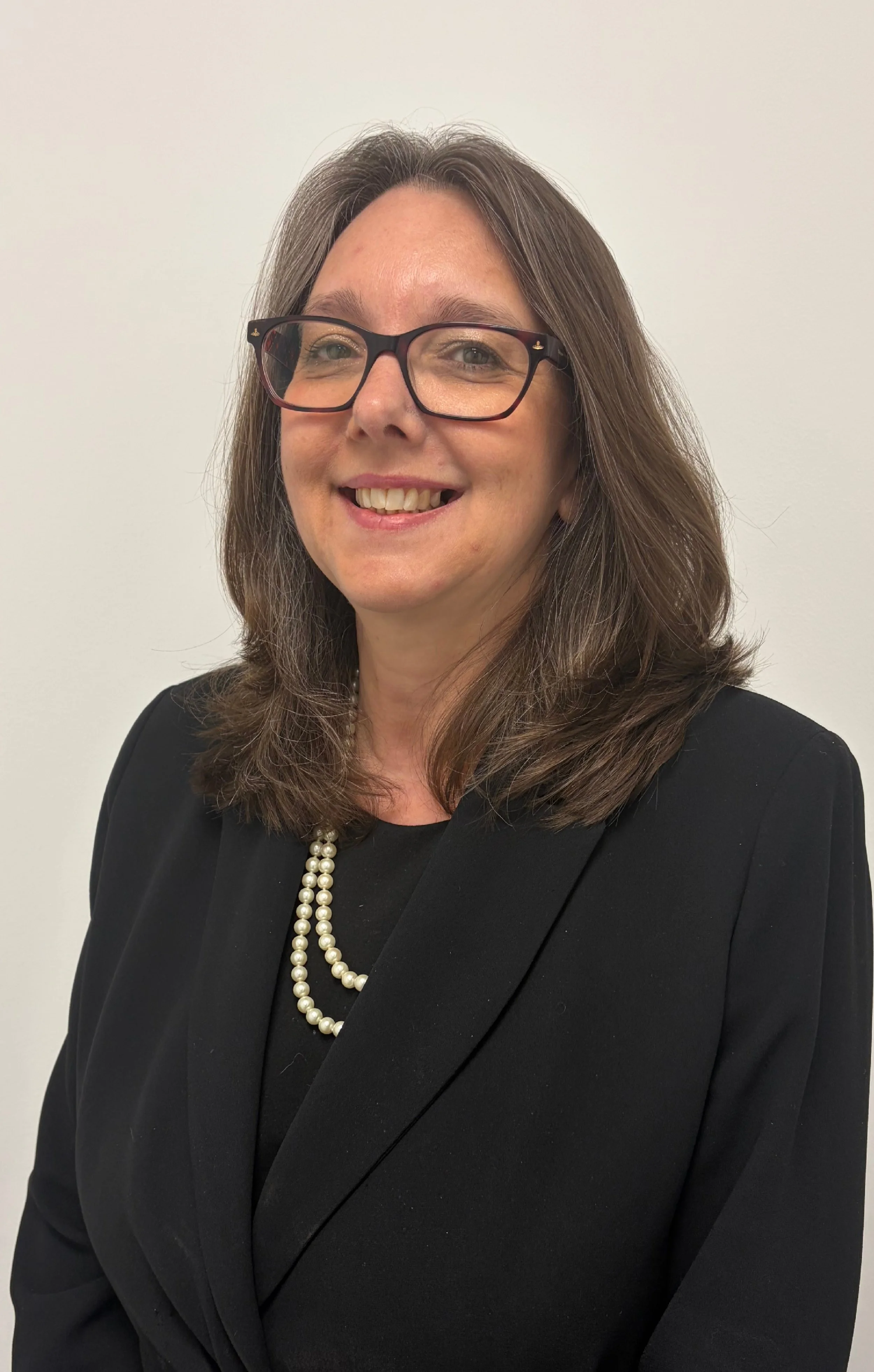A woman with shoulder-length brown hair, wearing glasses, a black blazer, and a colorful beaded necklace, smiling against a beige background.