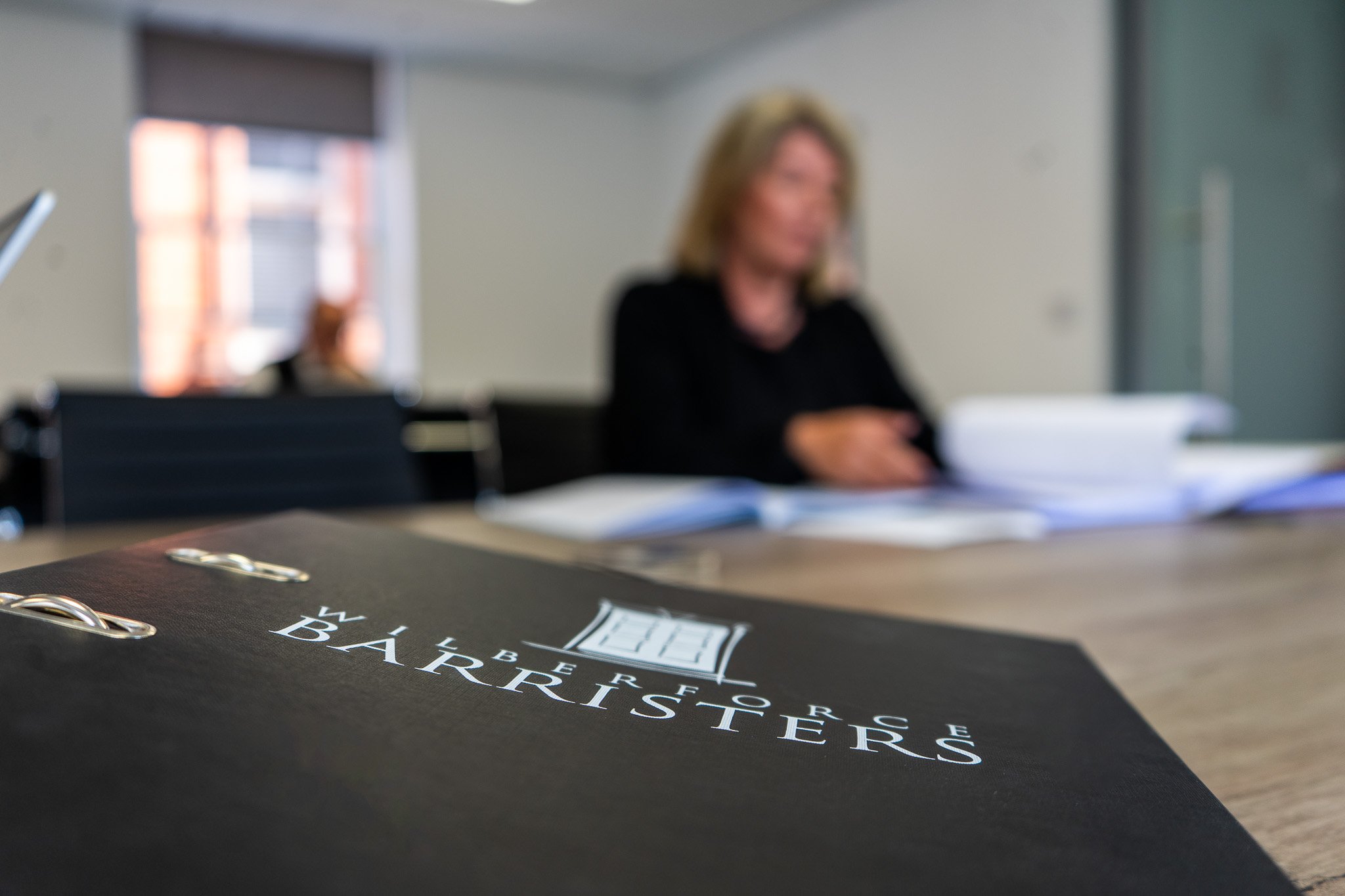 Close-up of a black binder with the words "WILBERFORCE LAWBARRIERS" printed on it, on a desk in a meeting room, with a woman in a black blazer sitting at the table in the background.