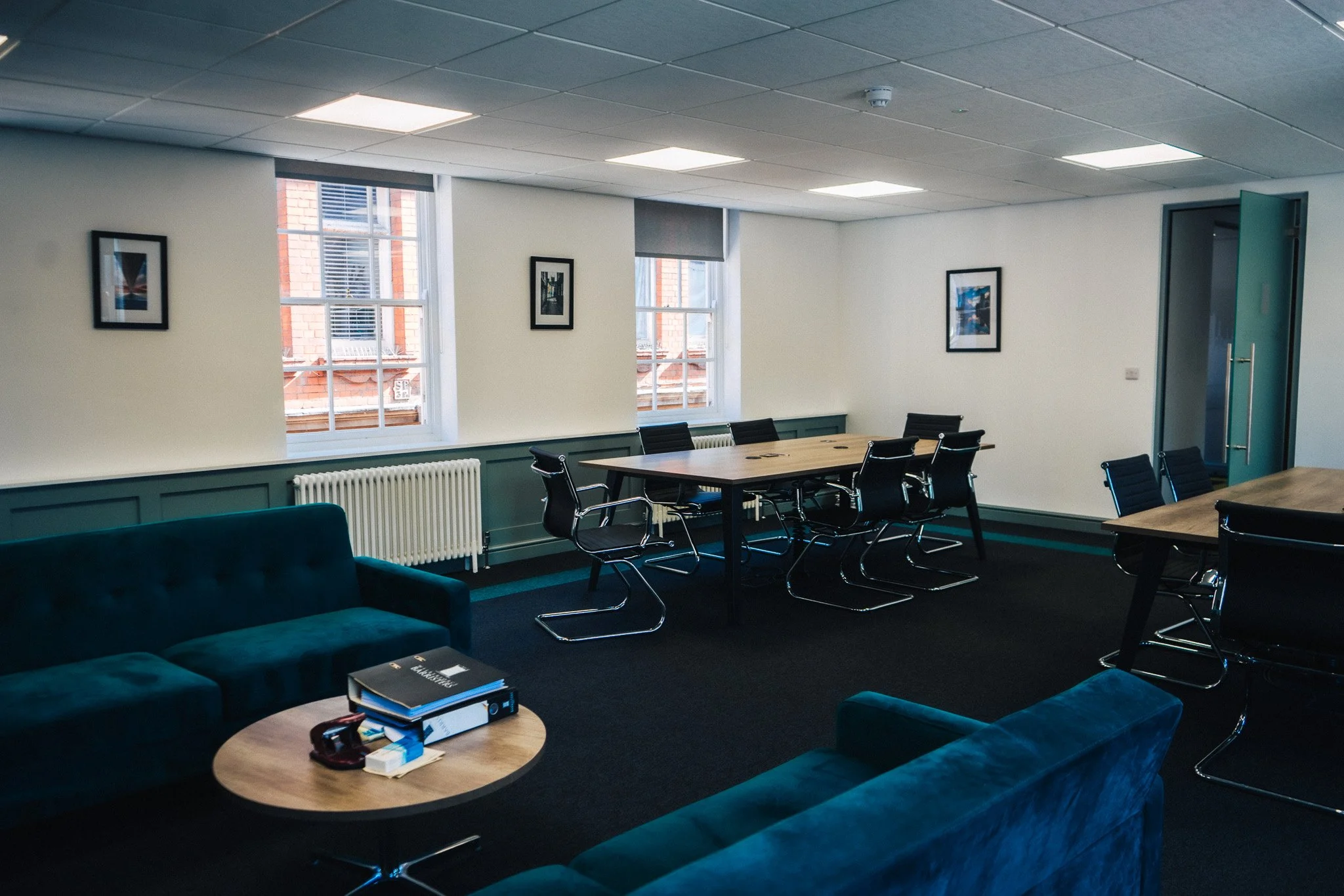 An office conference room with two teal sofas, a round wooden coffee table with binders and office supplies, a rectangular table with six black chairs, and two windows with framed pictures on the wall.