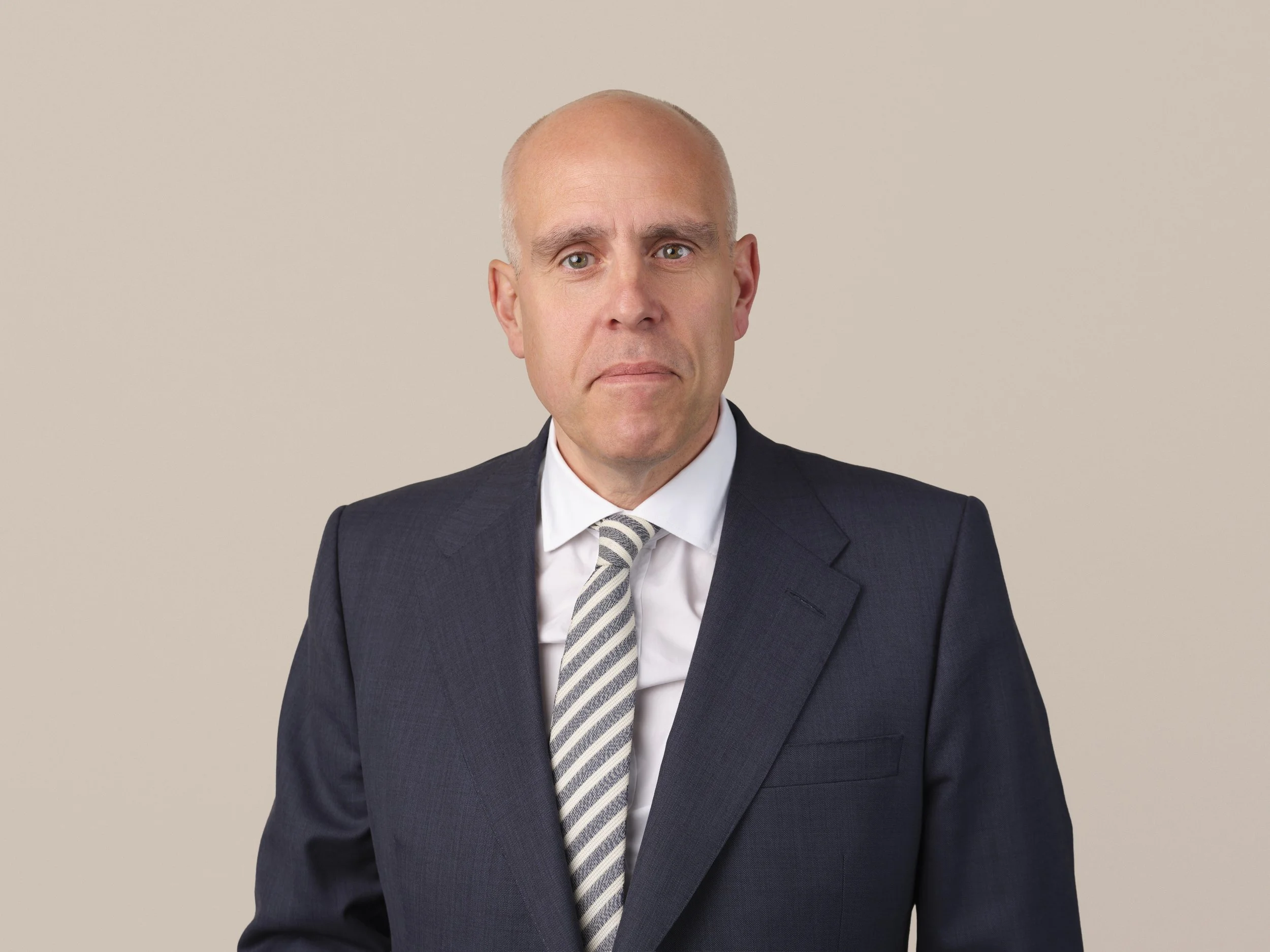 A middle-aged man with a serious expression, dressed in a navy blue suit, white shirt, and a navy tie with small red patterns, against a light beige background.