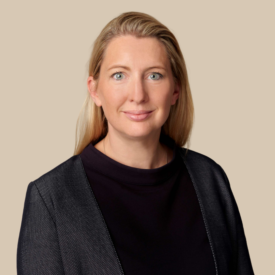 Portrait of a woman with blonde hair, blue eyes, wearing a black top and a blazer, smiling against a beige background.