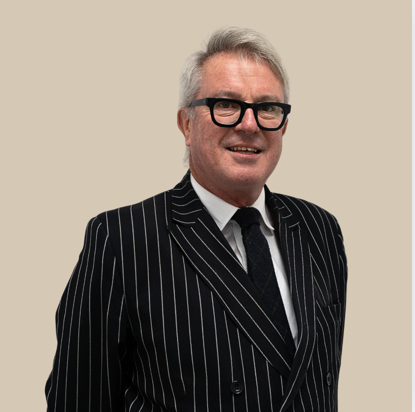 A middle-aged man with gray hair and glasses, wearing a black pinstripe suit and a white shirt, standing against a plain beige background.