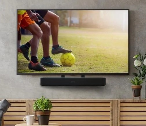 A TV displays a soccer game with players’ legs and a yellow ball; plants and a sonos beam soundbar are below the TV on a wooden bench.