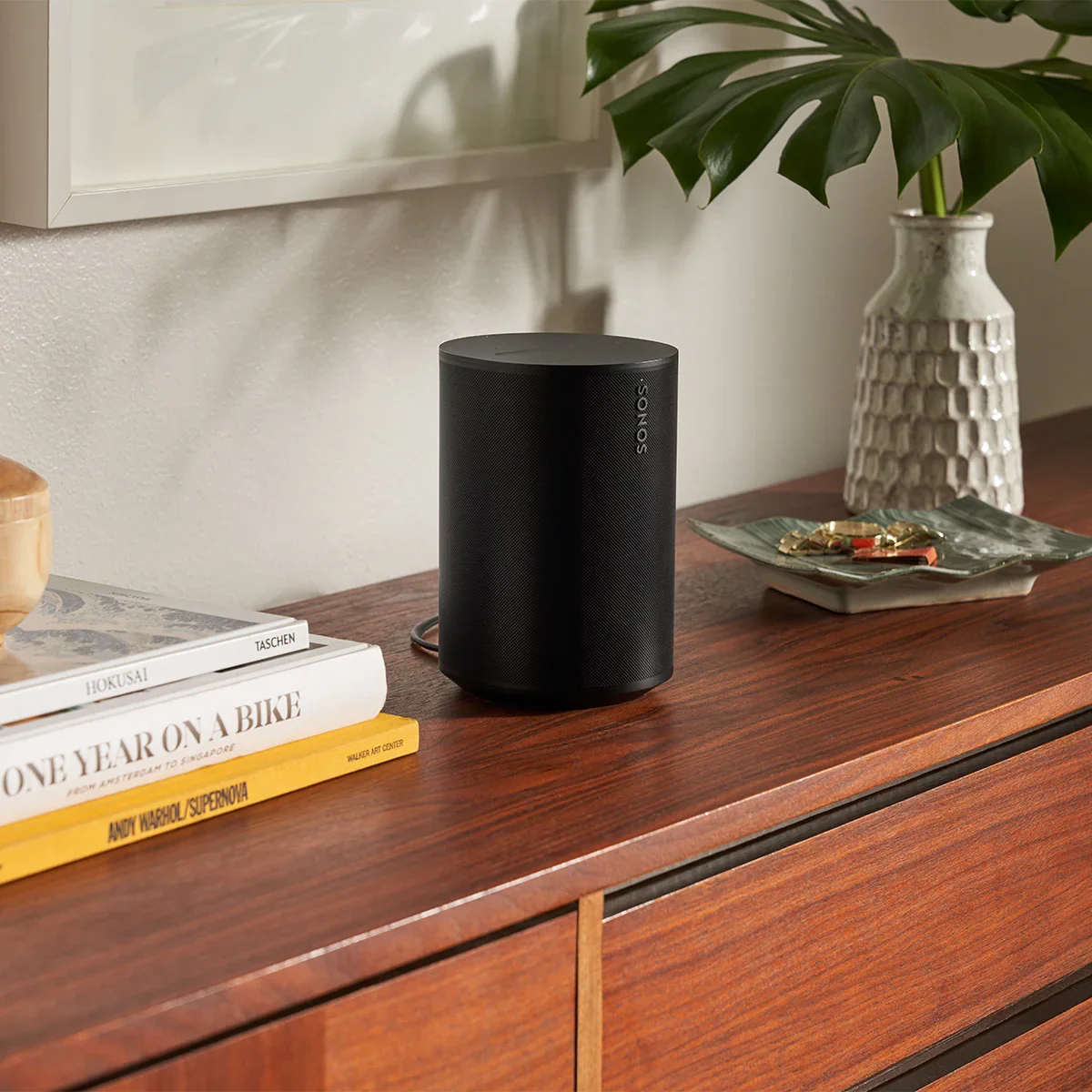 A black era 100 Sonos speaker sits on a wooden sideboard next to books, a plant, and a decorative tray.