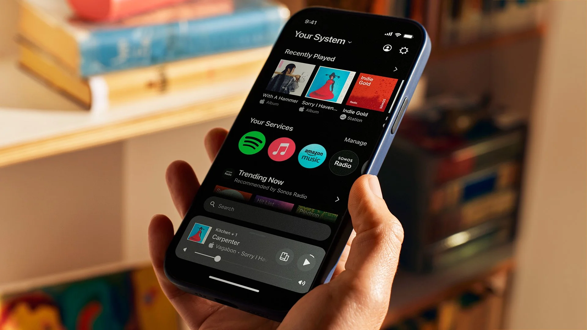 A hand holds a smartphone displaying a sonos music streaming app with playlists and music services on the screen.