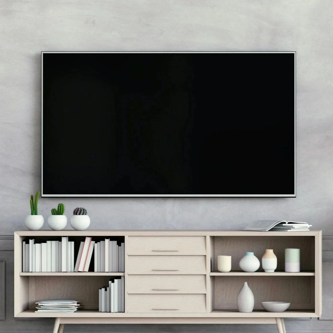 A wall-mounted flat-screen TV above a light wood cabinet with books, plants, and ceramic decor in a modern living room.