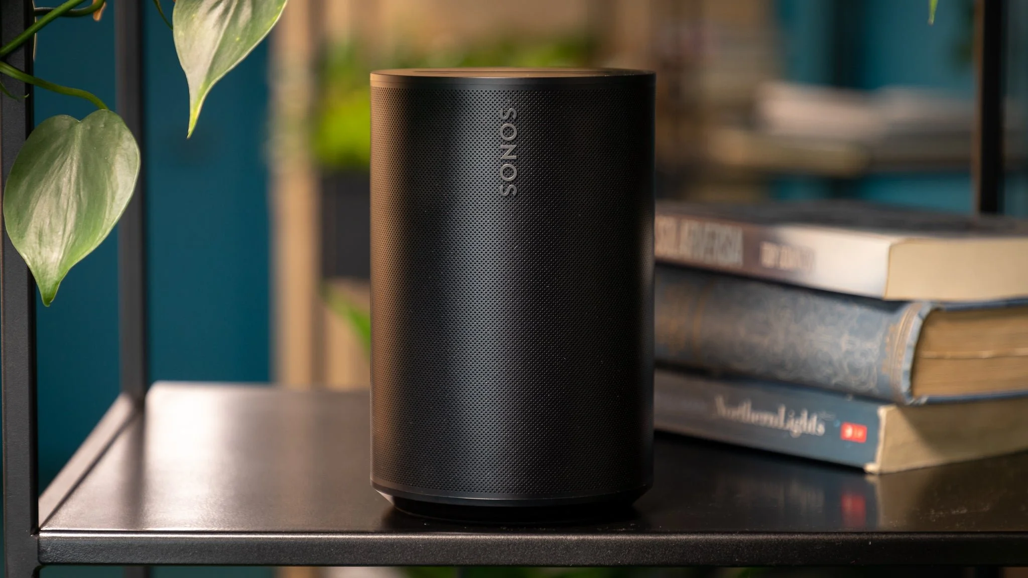 A black era 100 Sonos speaker sits on a shelf next to stacked books and a leafy plant.