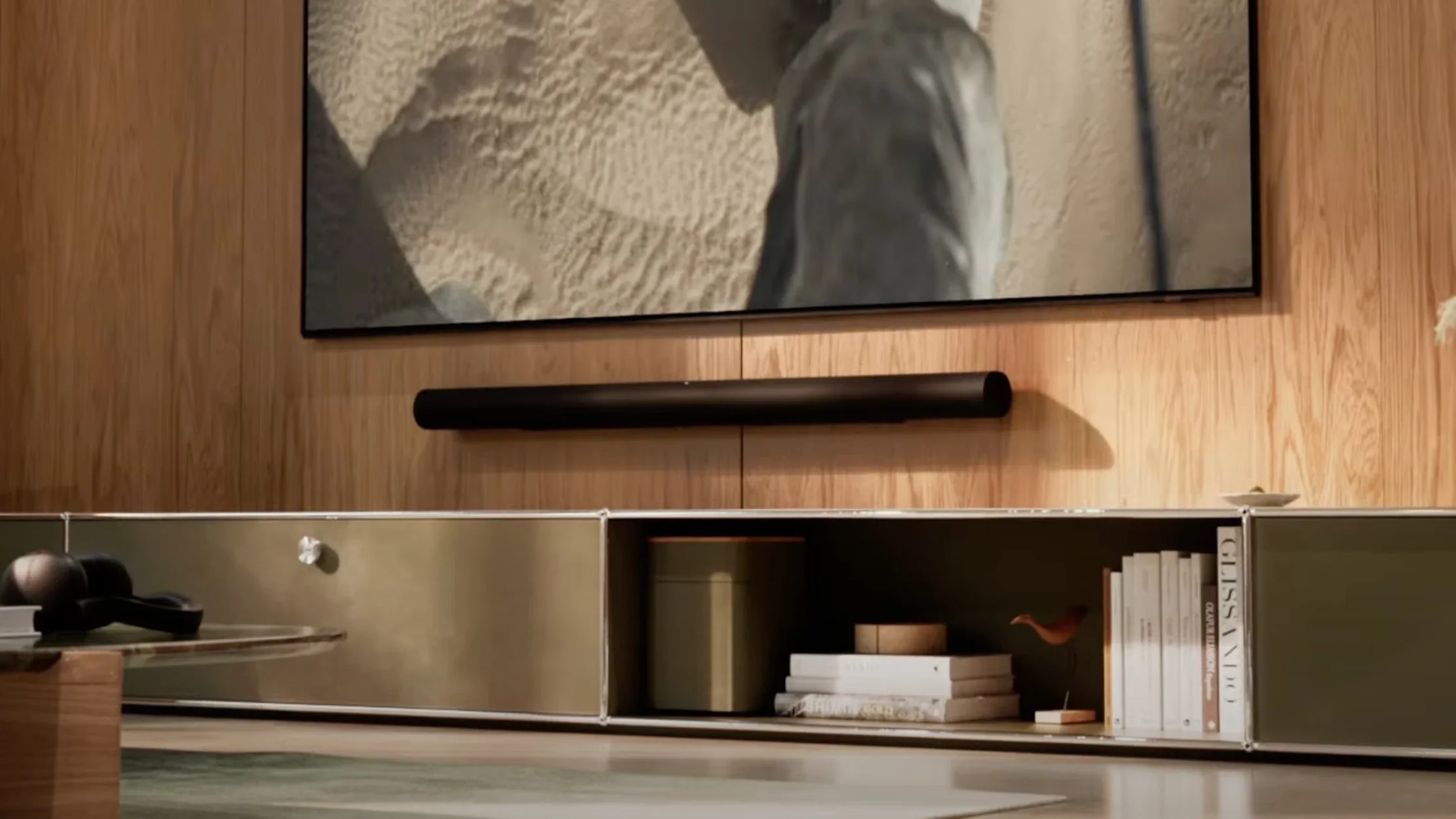 A modern TV on a wooden wall above a soundbar, with shelves below holding books, decor, and a lidded container.