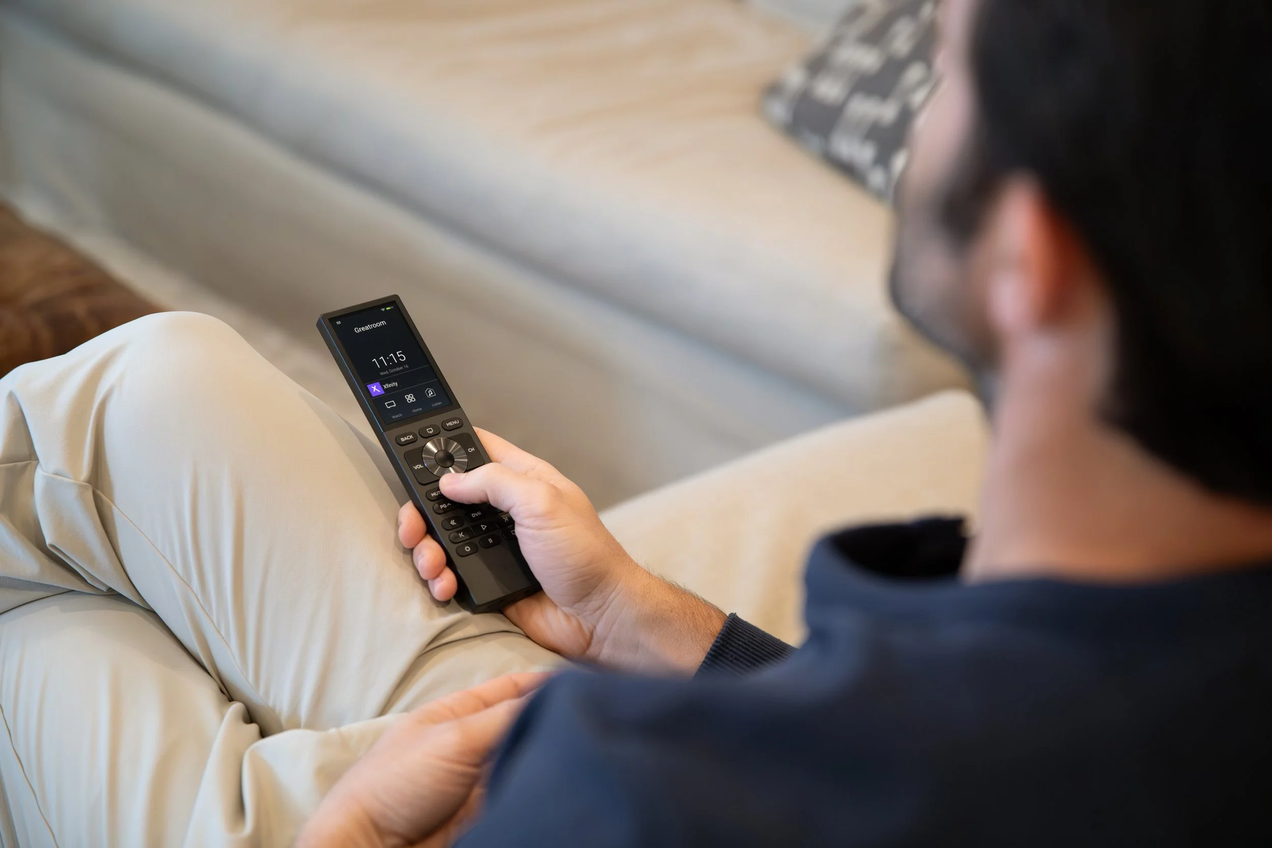 A man sitting on a beige couch holding a Home Cinema remote control with a screen displaying the time 11:15 and airport gate 'GreeceRoom' in an indoor setting.