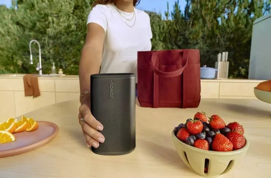 A woman sets a Sonos play portable speaker on a table outdoors with fruit and a red bag in the background.