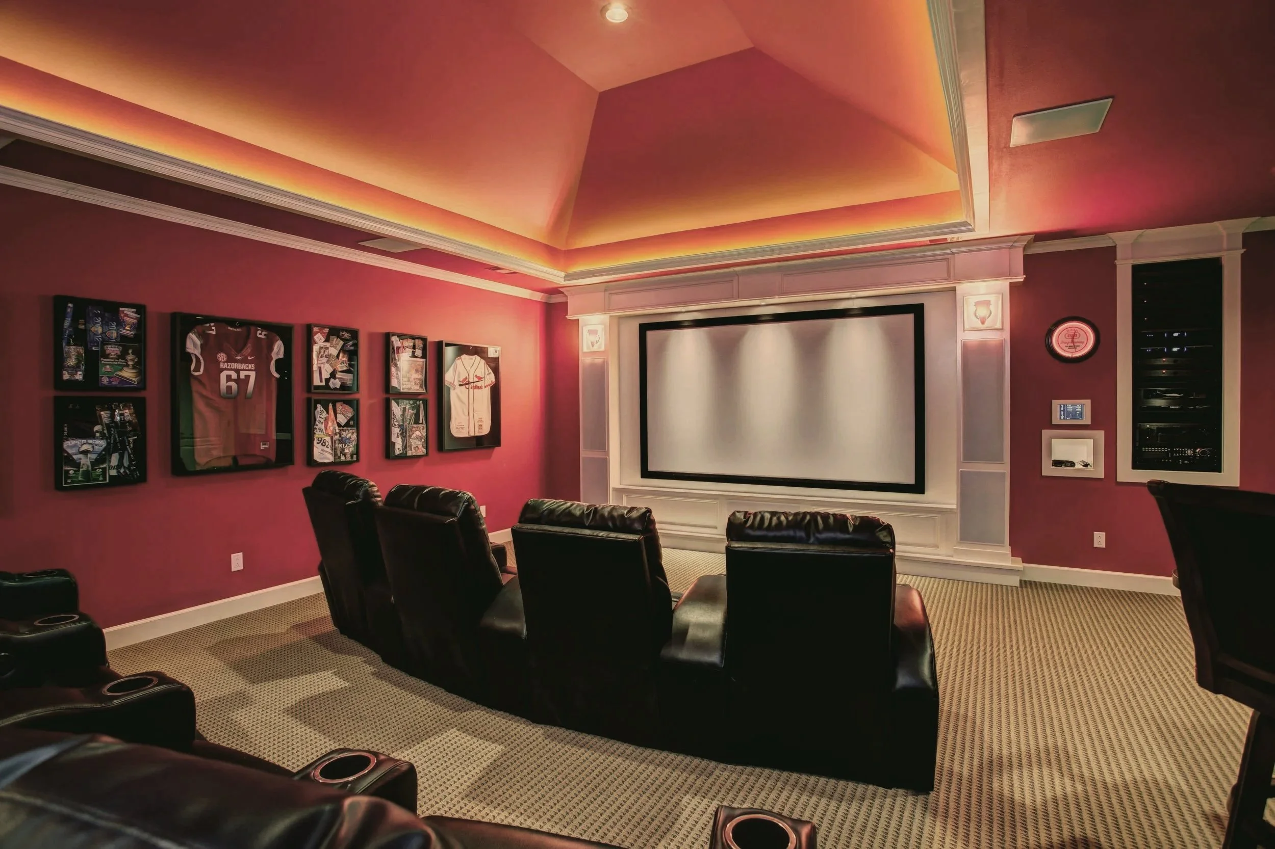 A home cinema room with a large projection screen, 5 black leather recliner chairs, framed sports jerseys and memorabilia on pink walls, and illuminated ceiling with warm lighting.