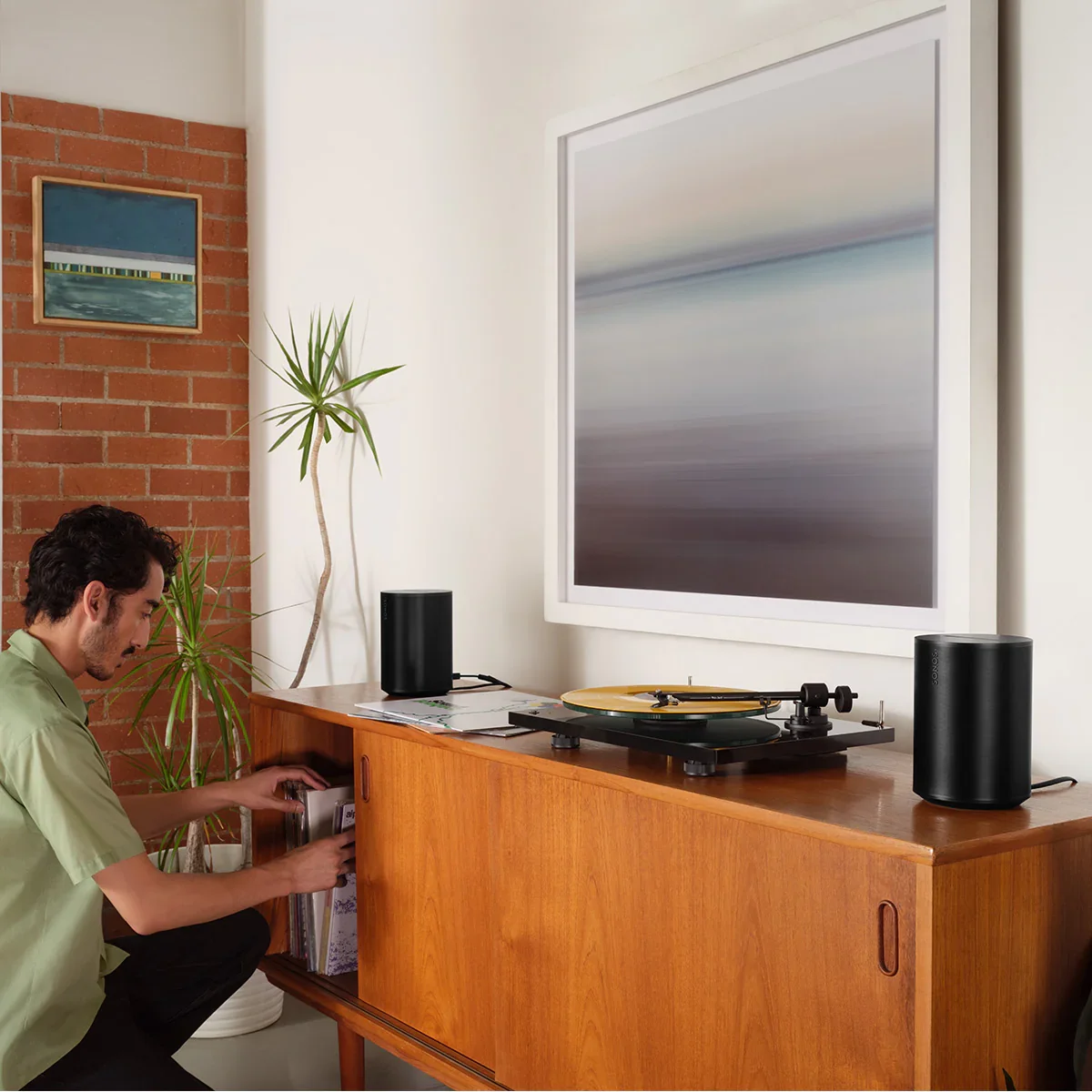 A person selects a vinyl record from a cabinet next to a turntable, sonos era 100 speakers, and modern art in a stylish living room.
