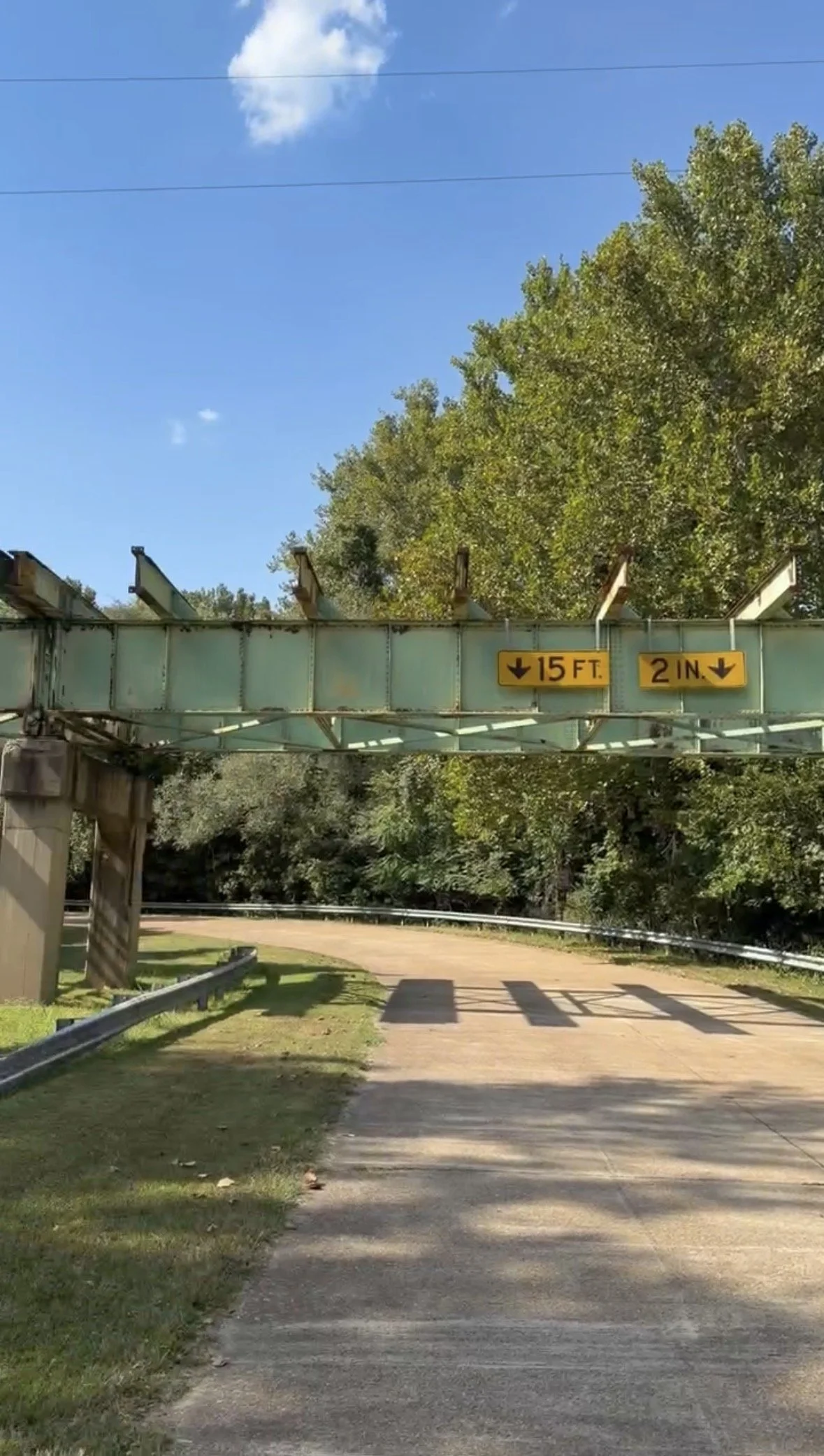 Blue sky over a dated green bridge with 2 yellow signs and a dirt road underneath