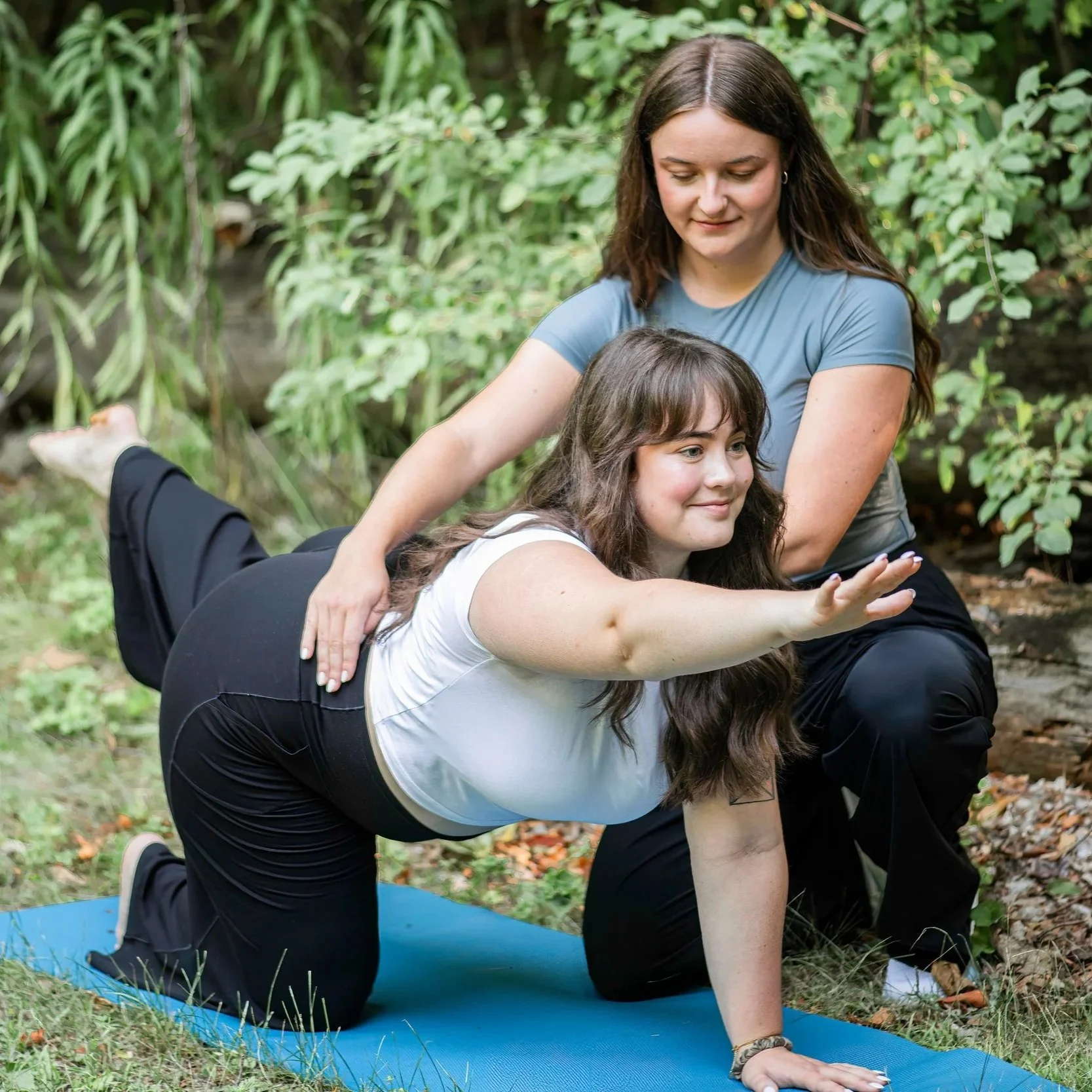 Two women practicing yoga outdoors on a blue mat, one helping the other into a pose with greenery in the background.