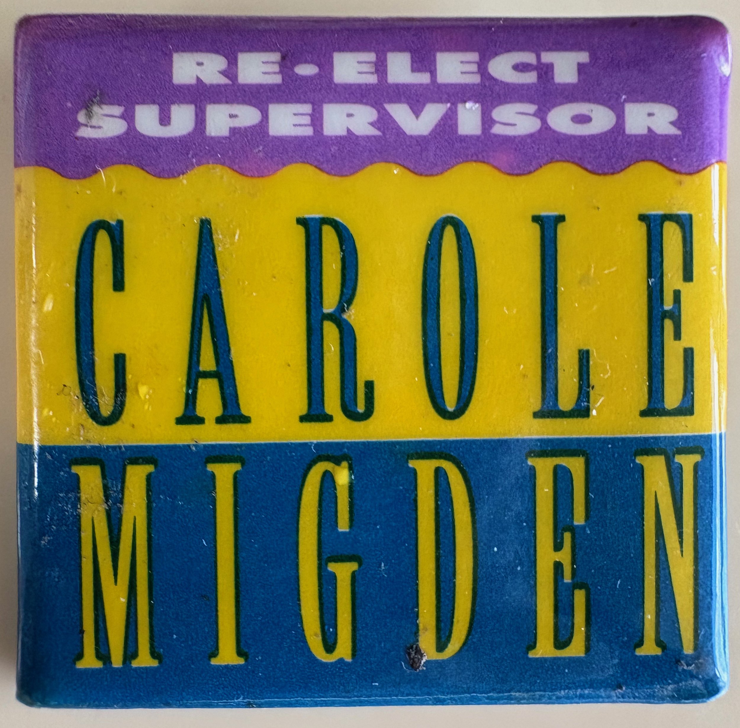 Colorful square sign with purple, yellow, and blue sections, reading 'Re-elect Supervisor Grace M. G. Den, 2024' in bold letters.