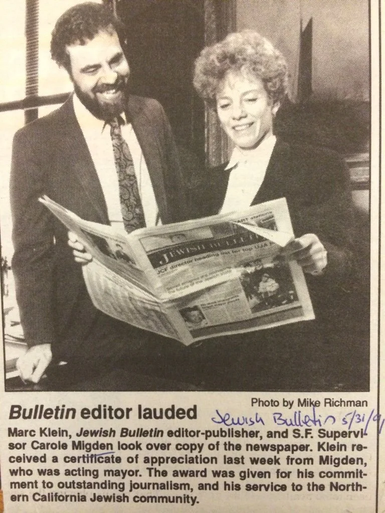 Two people, a man with a beard and a woman with curly hair, are looking at a newspaper and smiling. They are in an indoor setting, with a window and curtains behind them. The man is dressed in a suit and tie, and the woman is wearing a dark blazer ov