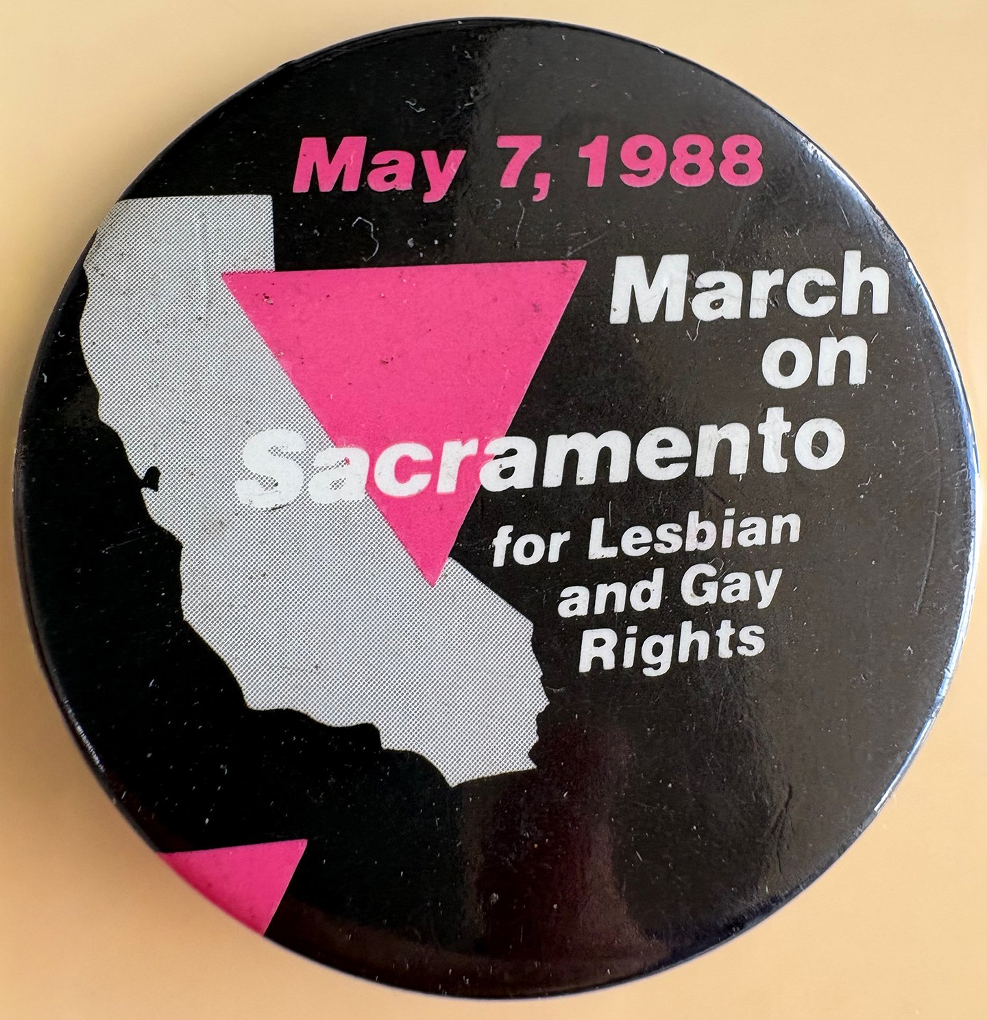 A button with a black background featuring a white silhouette of California, pink and white text, and pink and white geometric shapes celebrating the march on Sacramento for lesbian and gay rights on May 7, 1988.