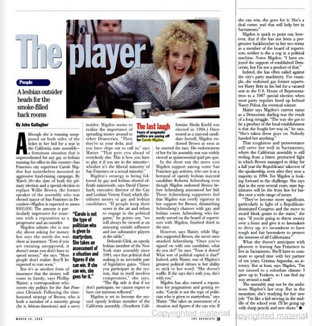 A woman with curly blonde hair smiling and speaking at a meeting. She is wearing a gray suit.