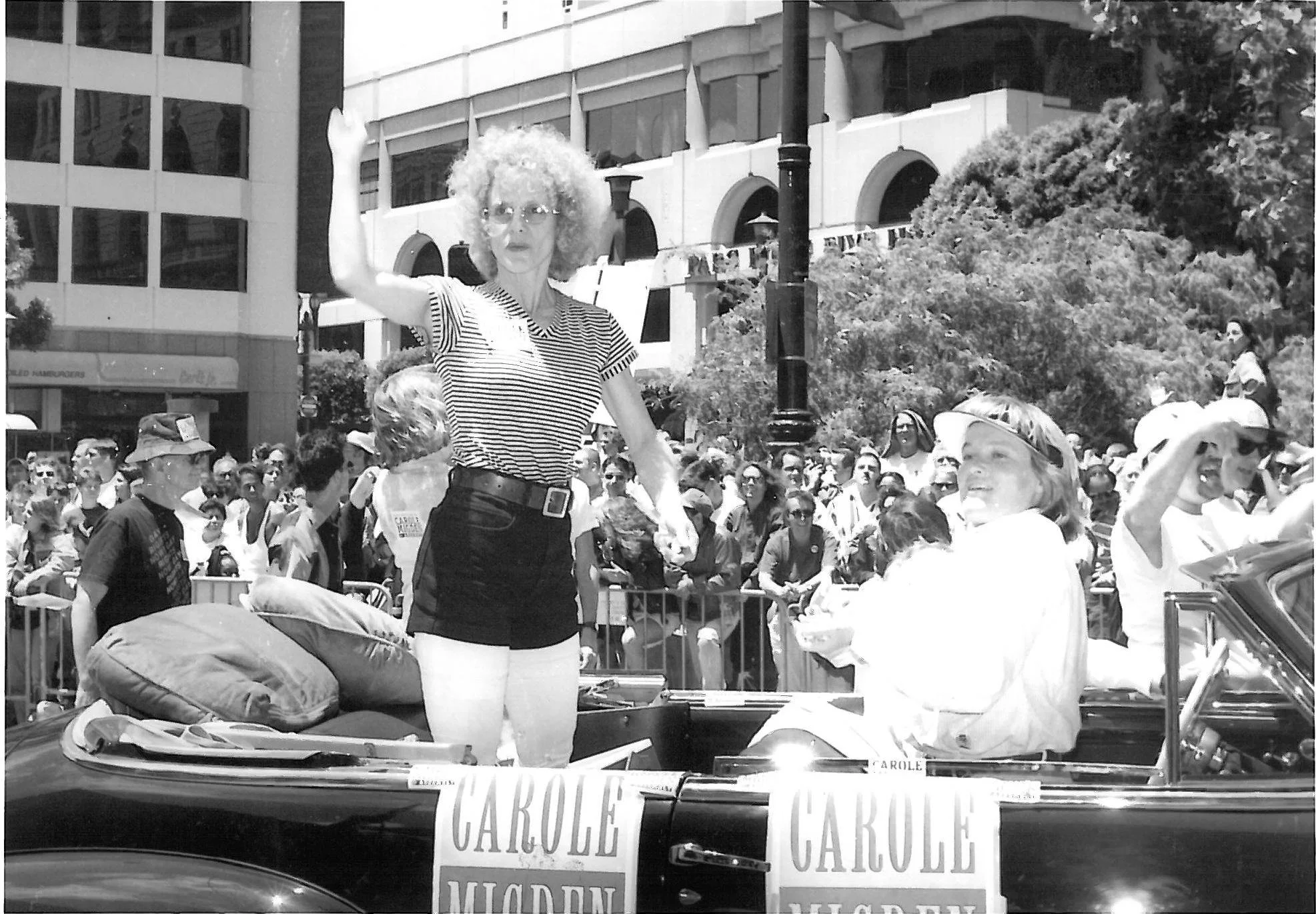 A woman with curly hair and glasses waving from the back of a car during a public event, with a crowd and city buildings in the background.
