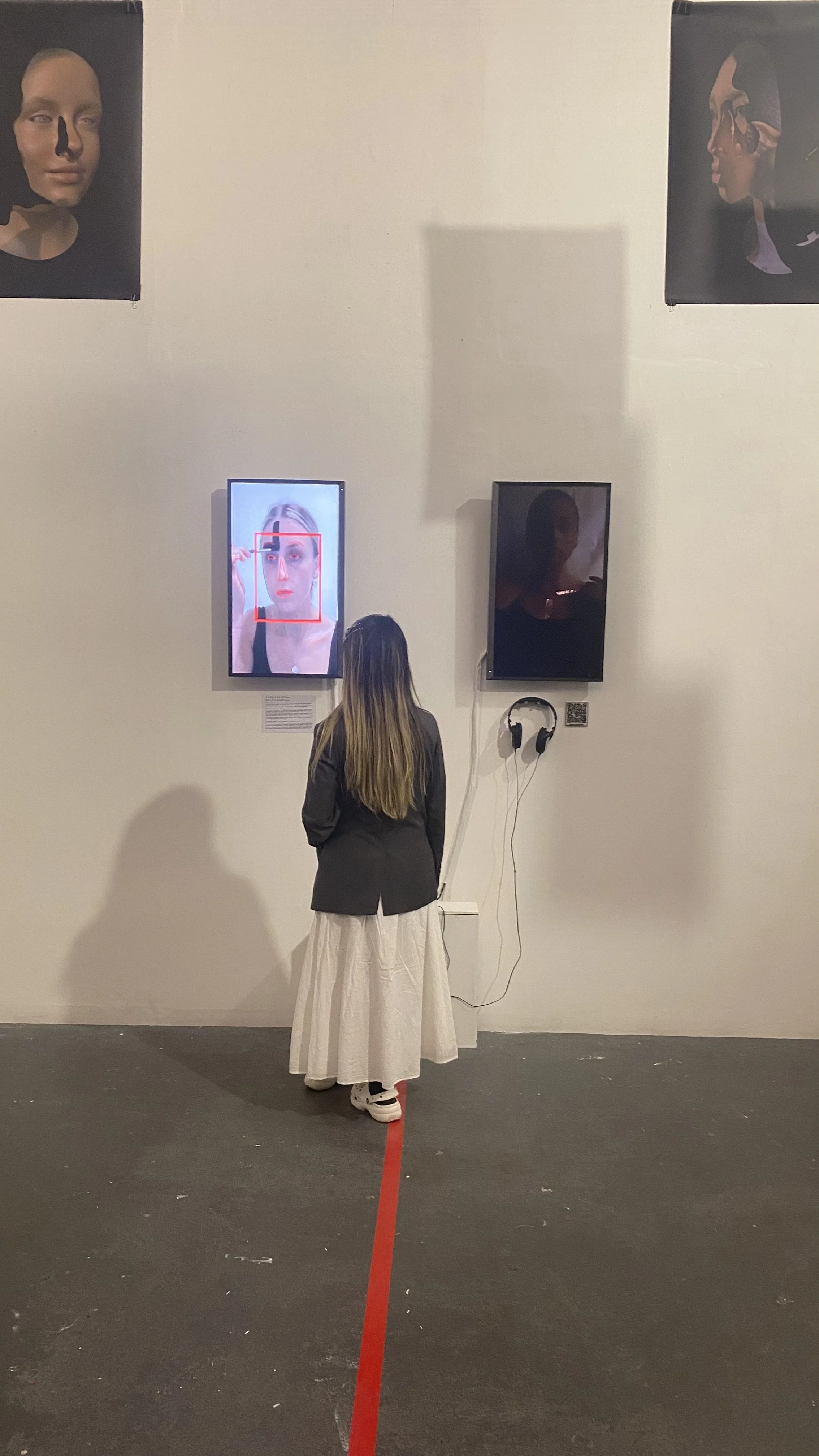 A woman with long hair standing in an art gallery, looking at a digital screen displaying a woman's face with a red square around her eye, with headphones hanging on the wall next to her. Large artistic portraits are on the wall above.