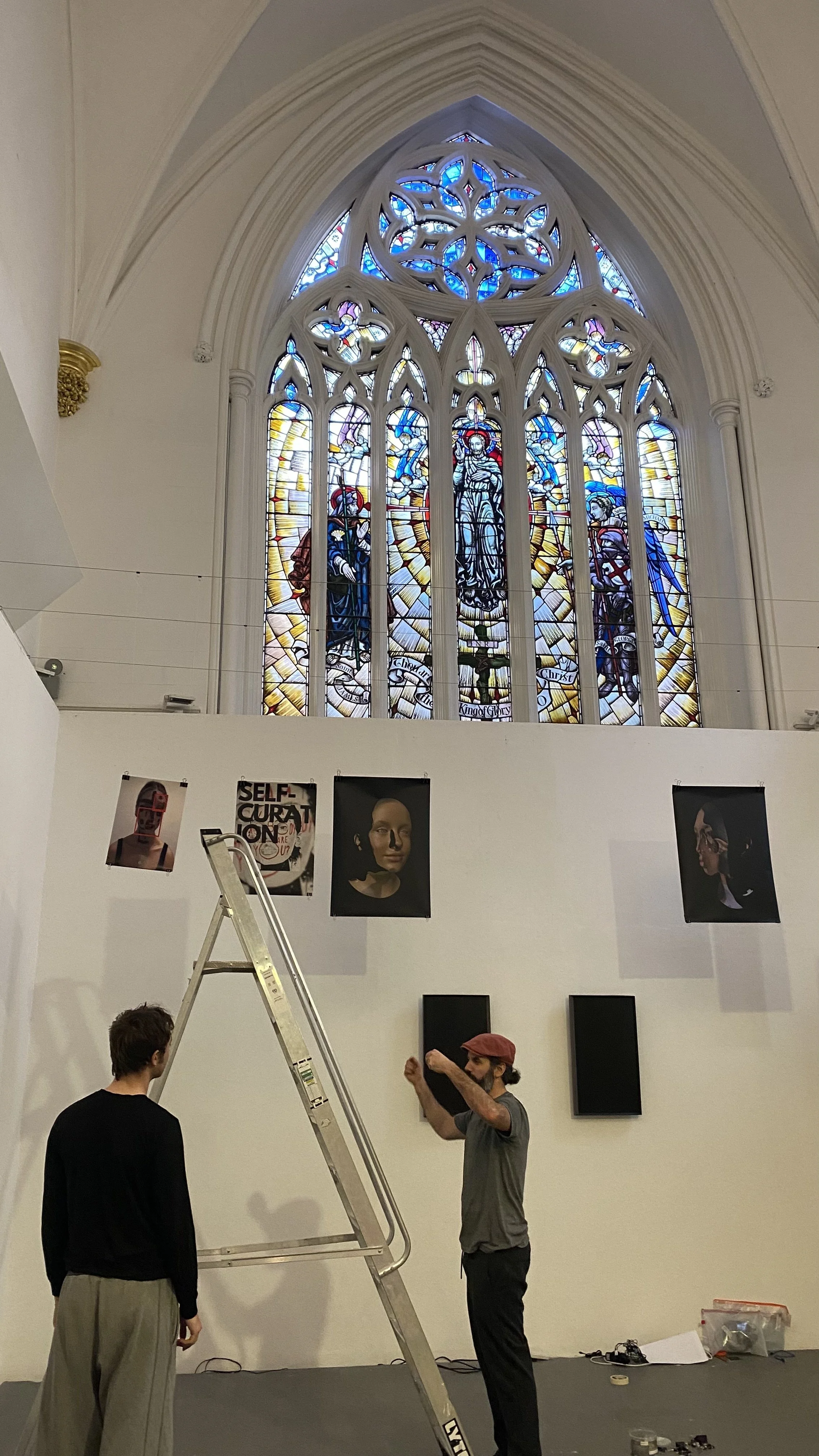 A stained glass window in a church with people setting up art exhibition below.