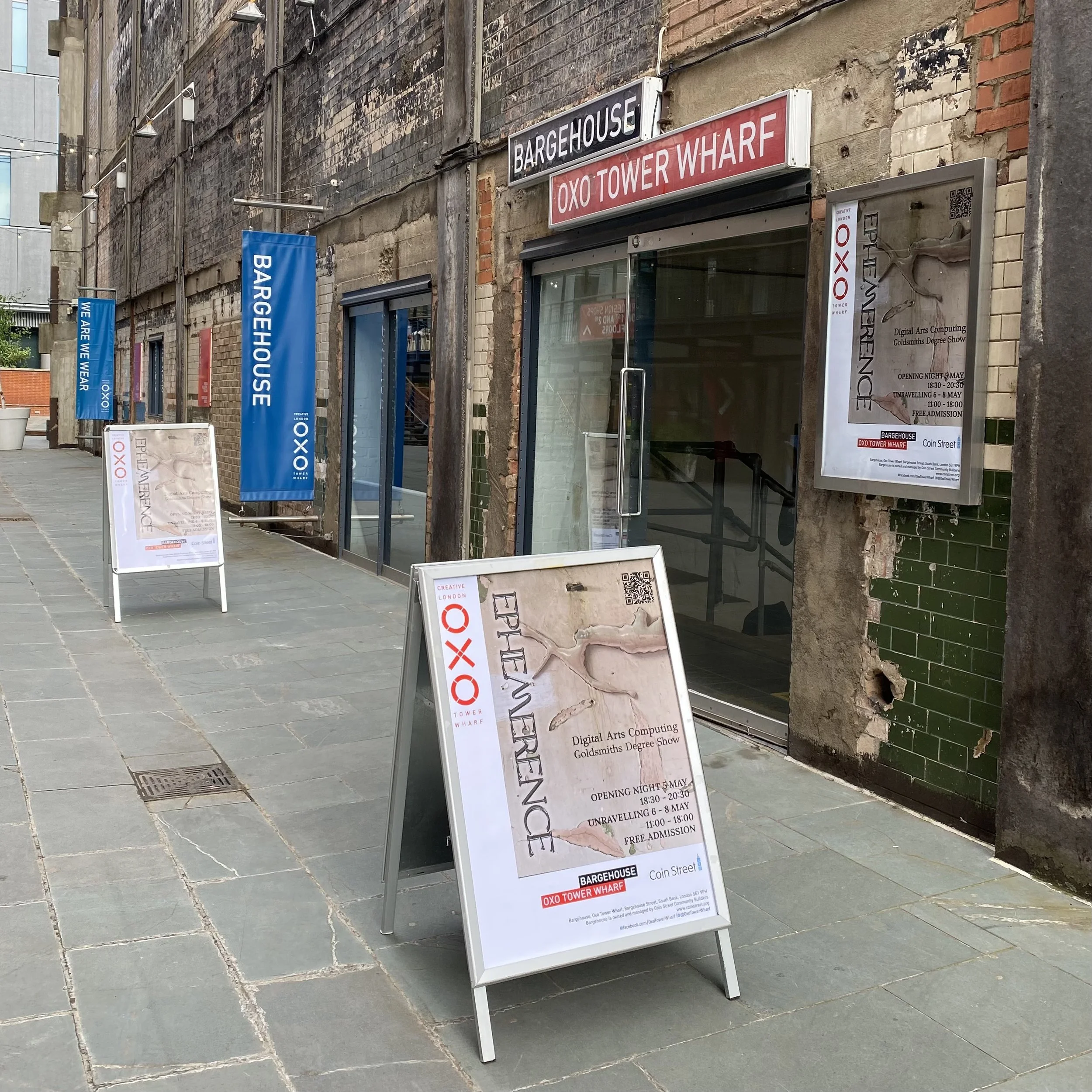 Street view of Oxo Tower Wharf, with an exhibition poster in front of it with details about opening night, hours, and admission.