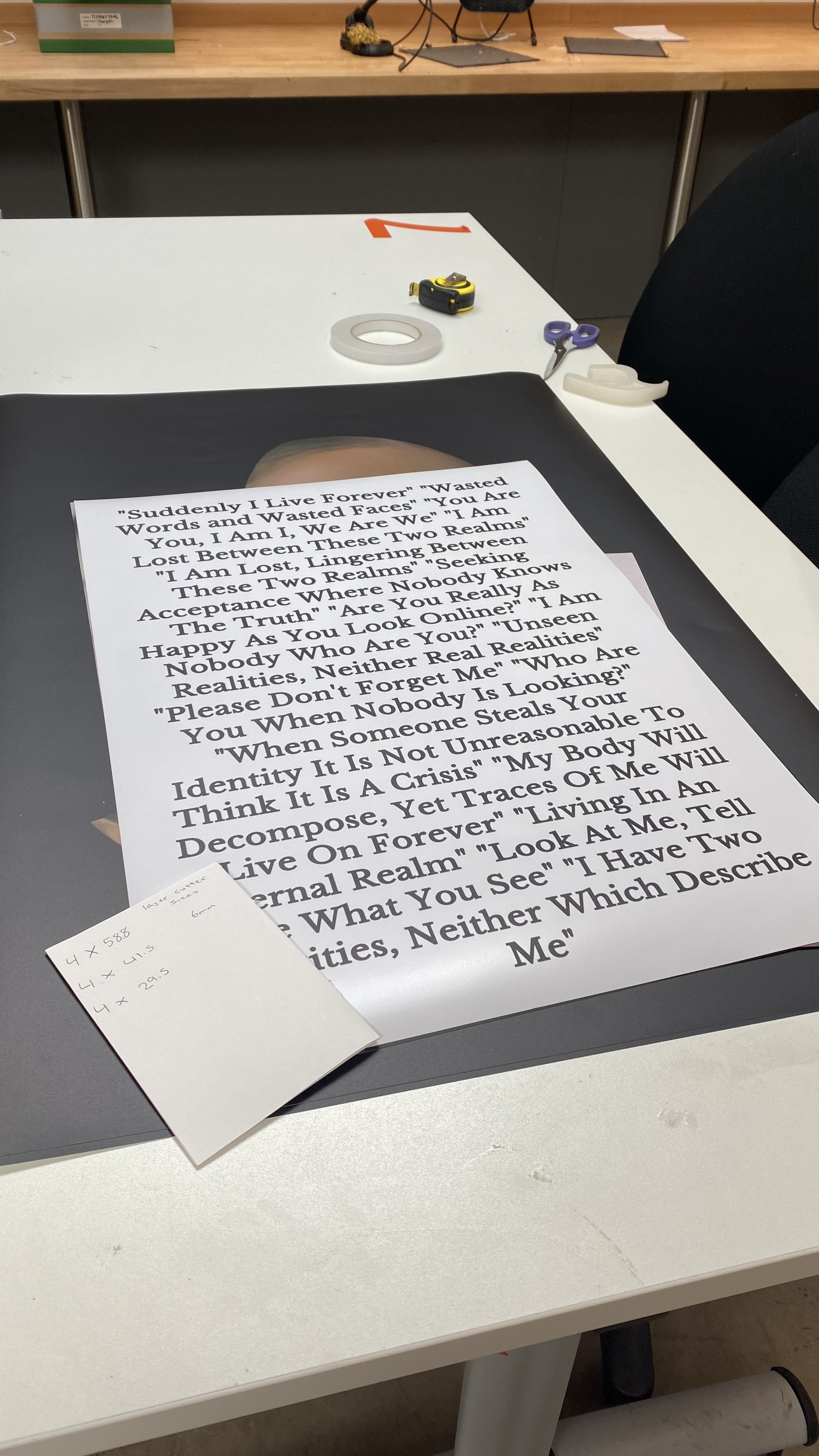 A printed sheet of paper with the lyrics of a song, placed on a work table, along with a small sticky note with handwritten measurements.
