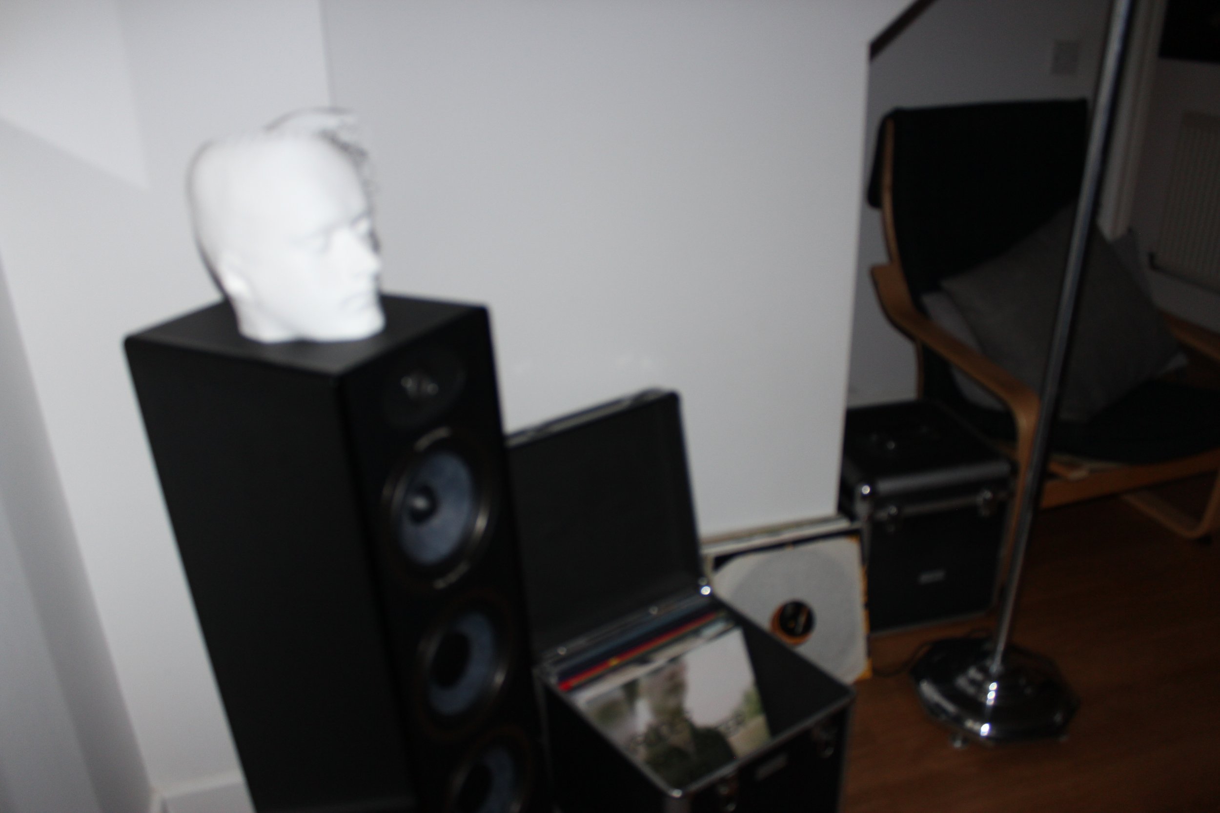 A white sculpture of a human head placed on top of a black speaker, with a chair and a mirror visible in the background.