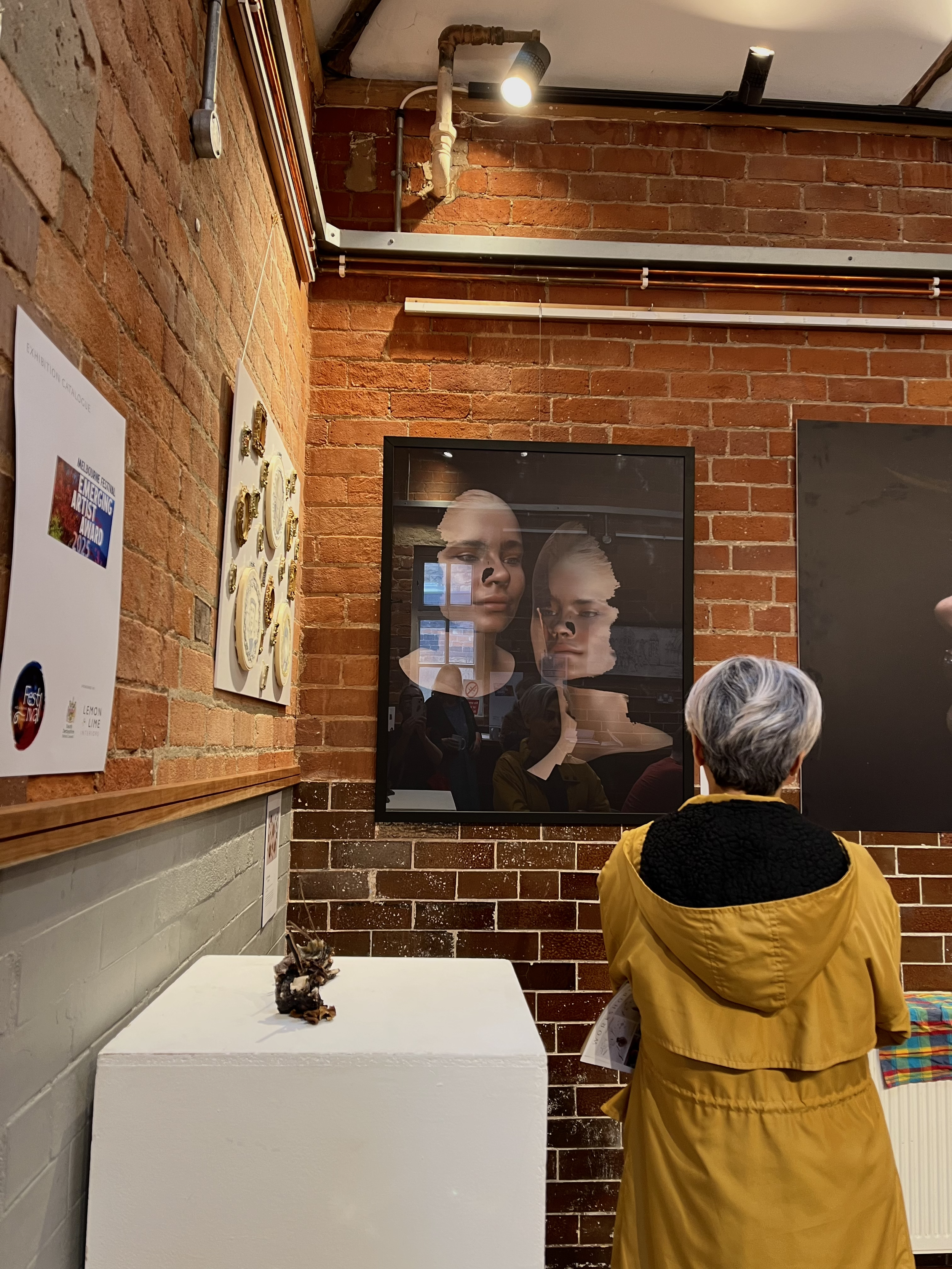 A woman with short grey hair wearing a yellow jacket stands in front of a portrait of two faces in a gallery with exposed brick walls.