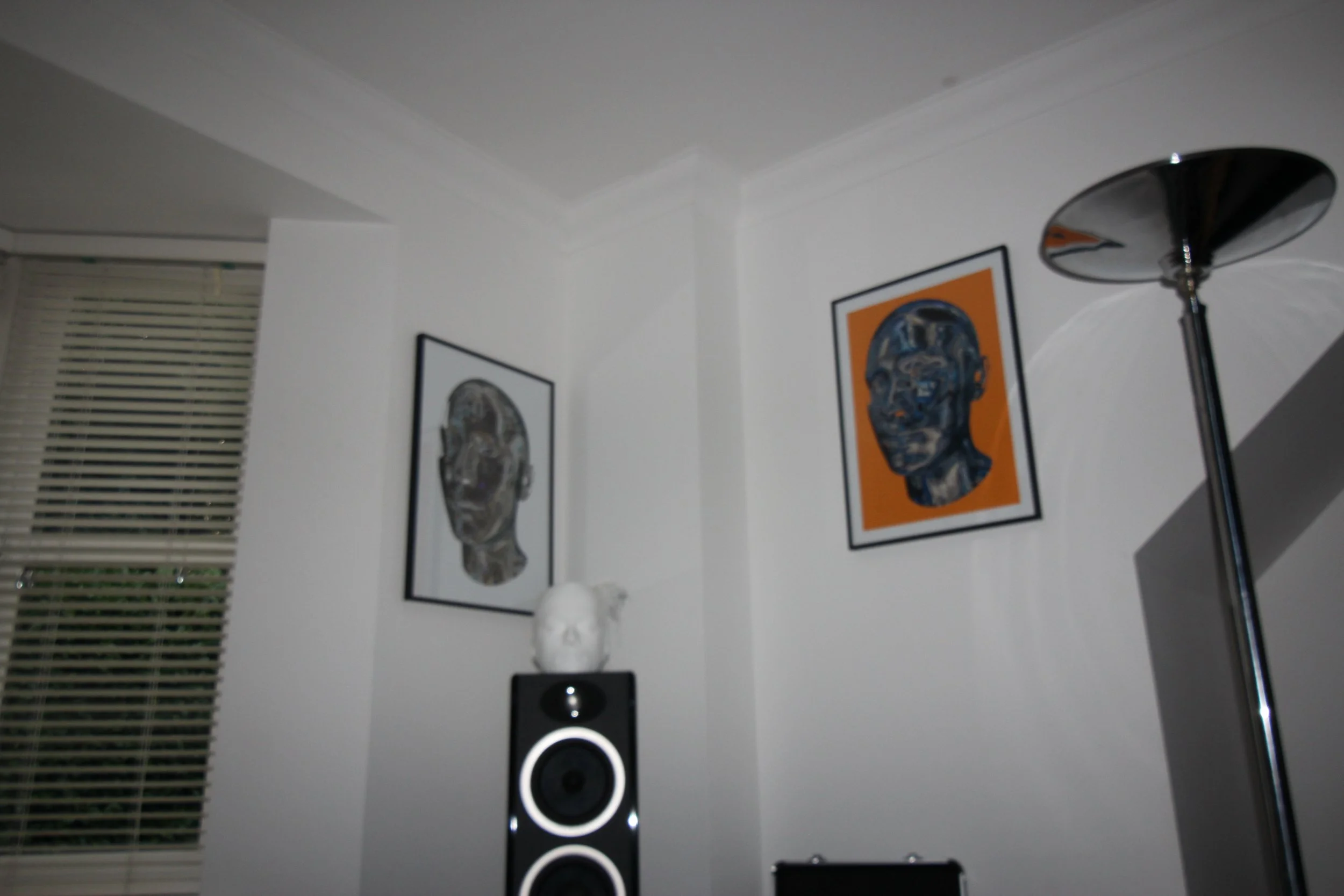 A corner of a room with framed posters of robotic heads hanging on the wall, a tall modern floor lamp, and a black speaker with white circles in front of the posters.