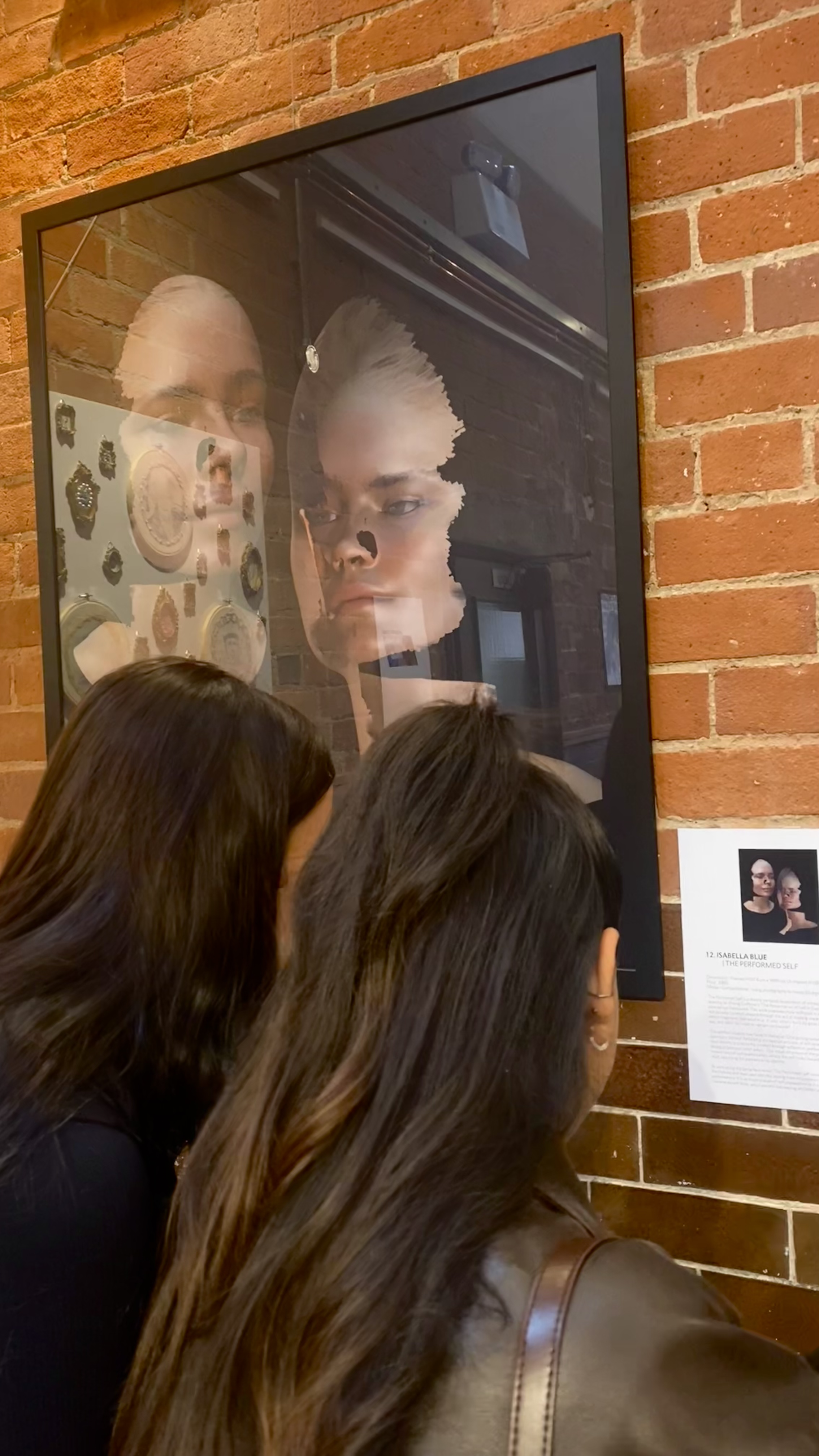 Women with long dark hair view an art piece titled 'The Performed Self' which is a large portrait.