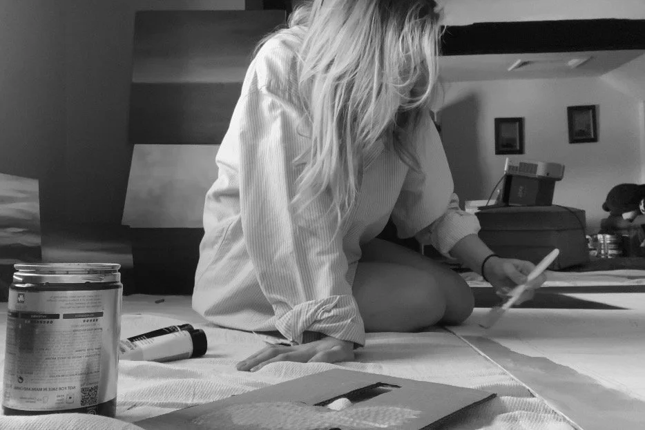 A woman with long hair covering the side of her face sitting on the floor painting on a large canvas.